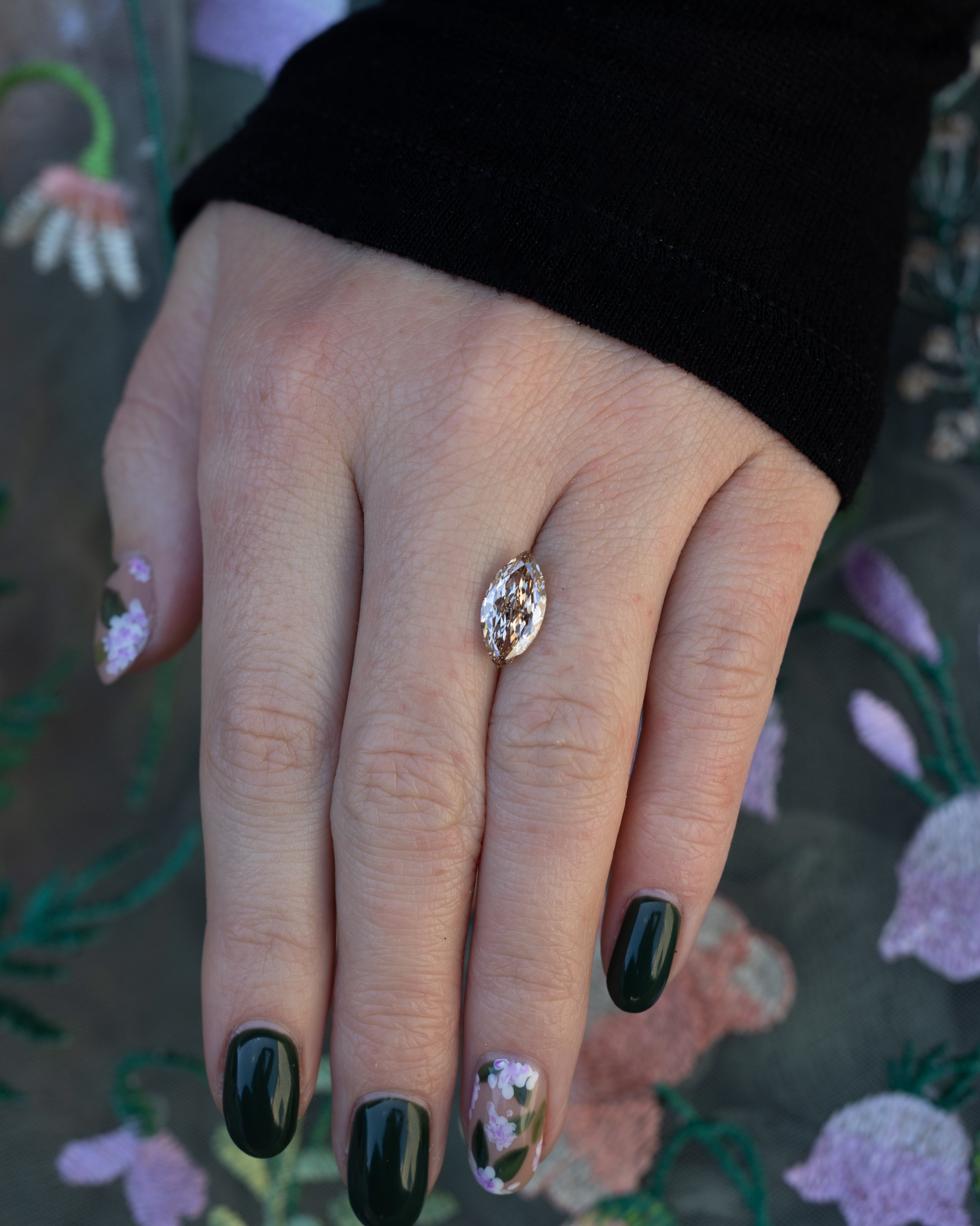 Hand holding a marquise champagne diamond with a black sleeve against a floral background.