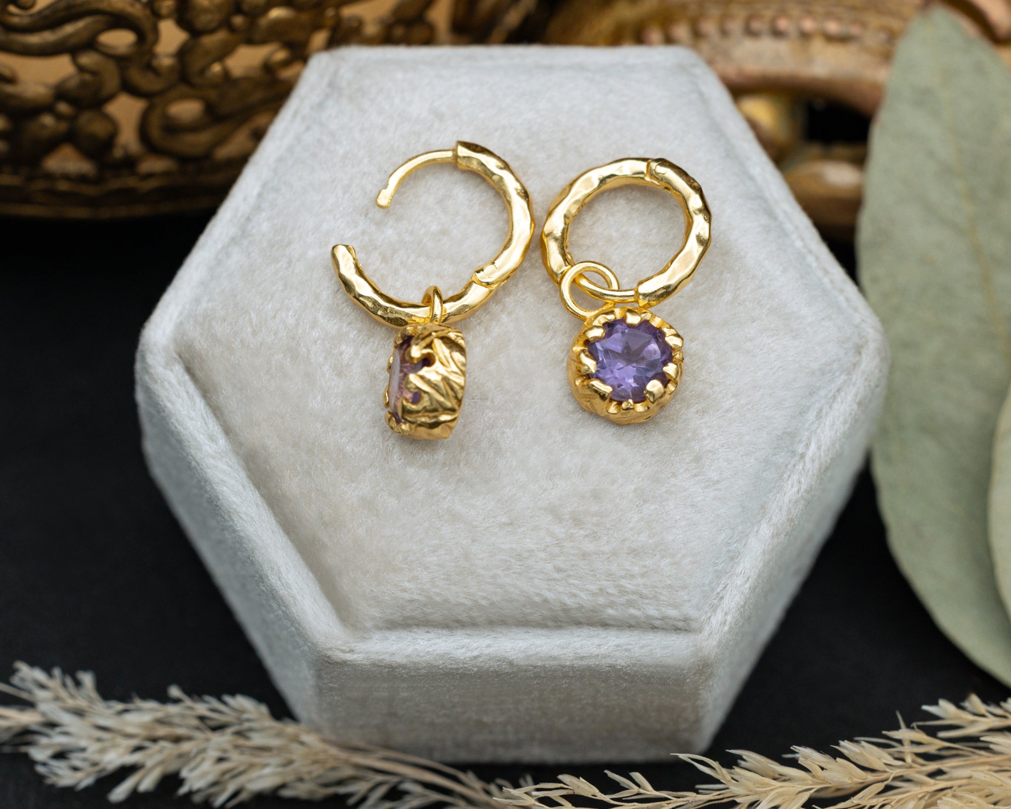 Gold hoop earrings with a purple amethyst on a hexagonal white jewelry box.