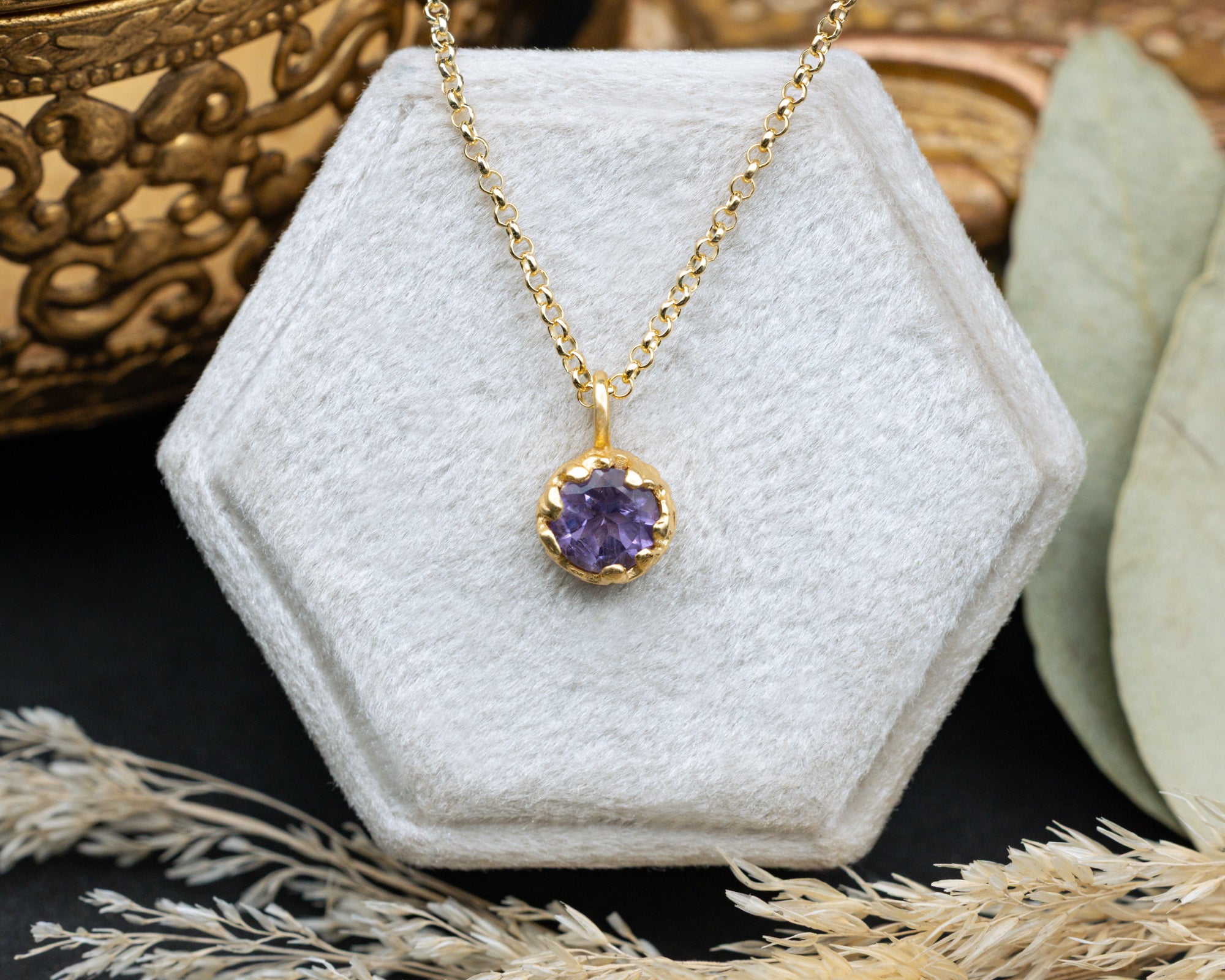 Gold necklace with a purple amethyst on a white hexagonal stand.