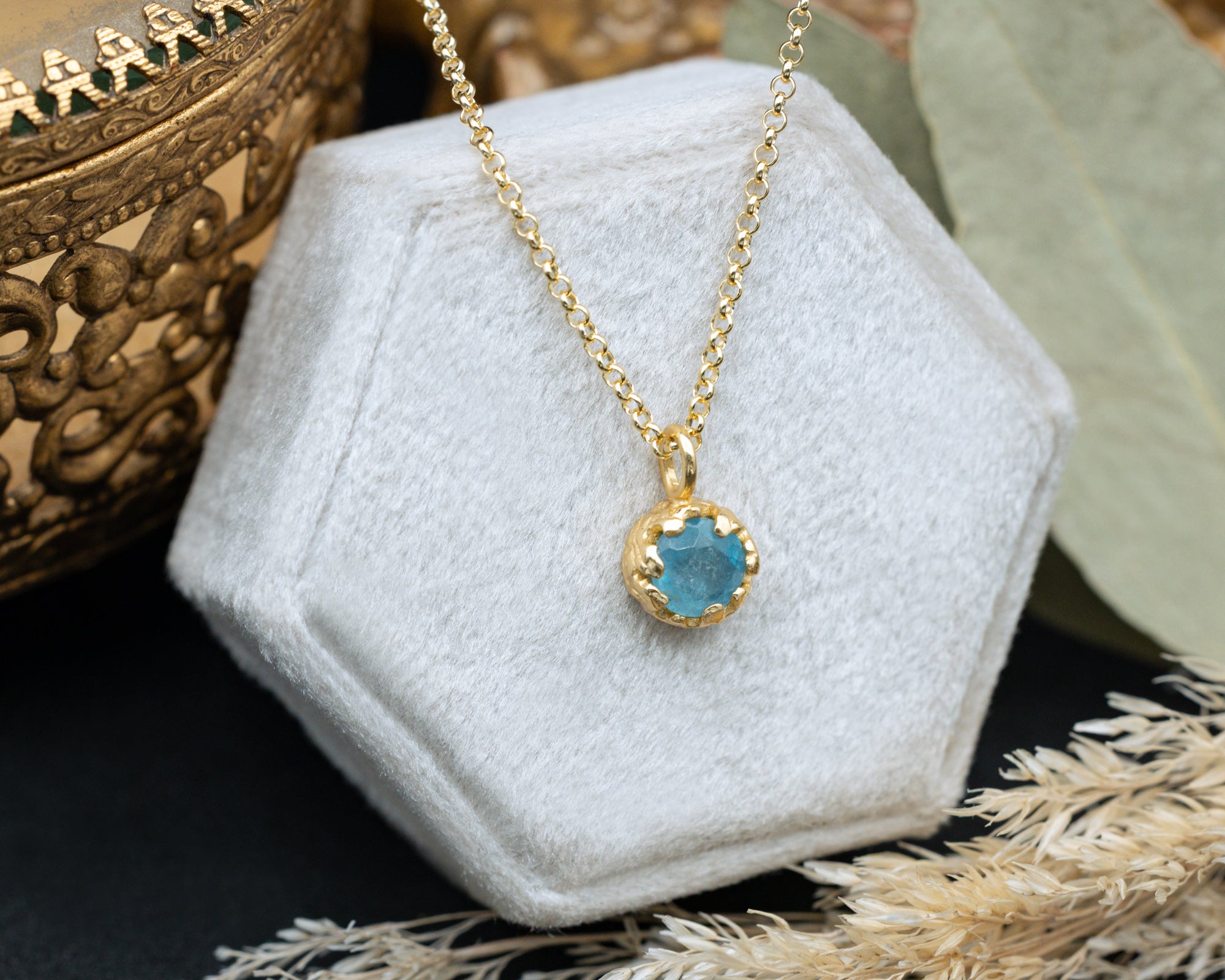 Gold necklace with a blue aquamarine on a white hexagonal stand.