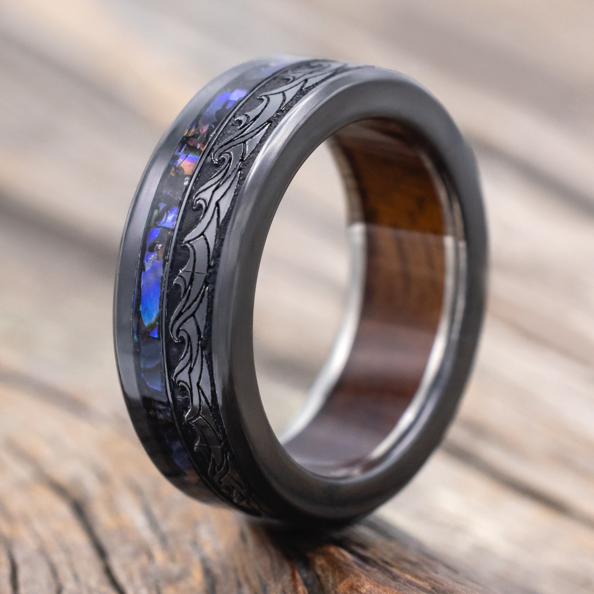 Black ring with paua shell, koa wood and wave engravings on a wooden surface