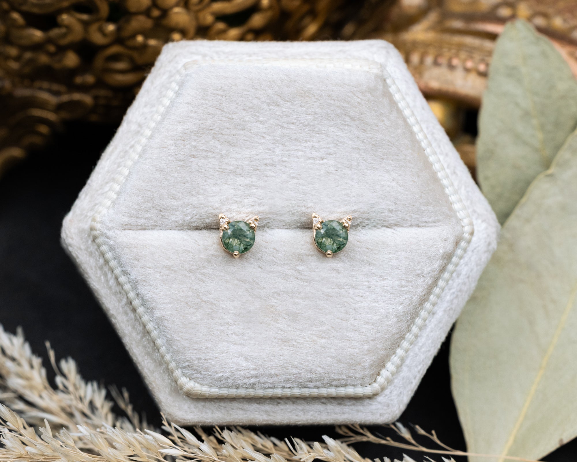 Pair of moss agate cat-shaped stud earrings on a white cushion against a decorative background
