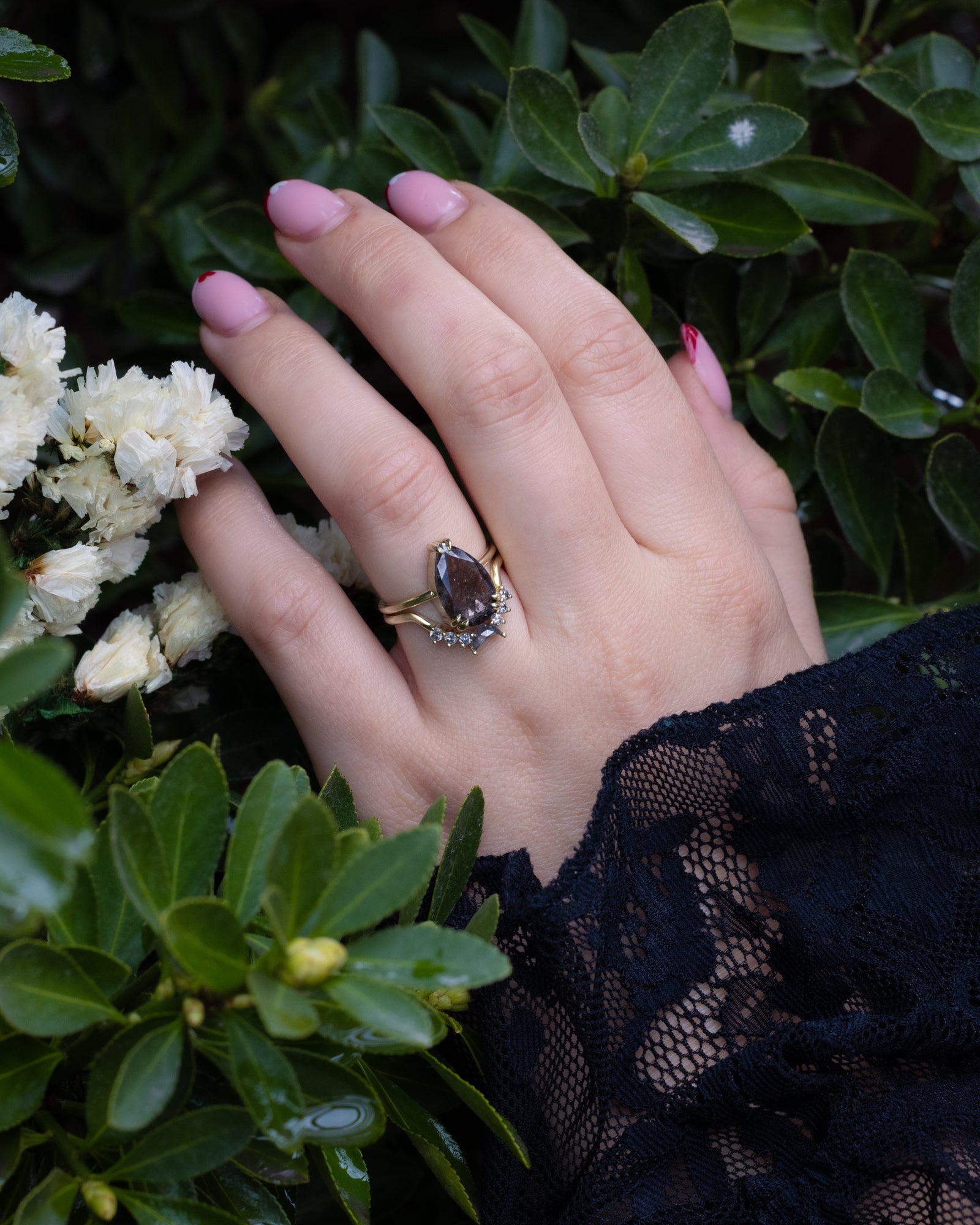 Hand wearing a ring with a salt & pepper diamond, against a green leafy background