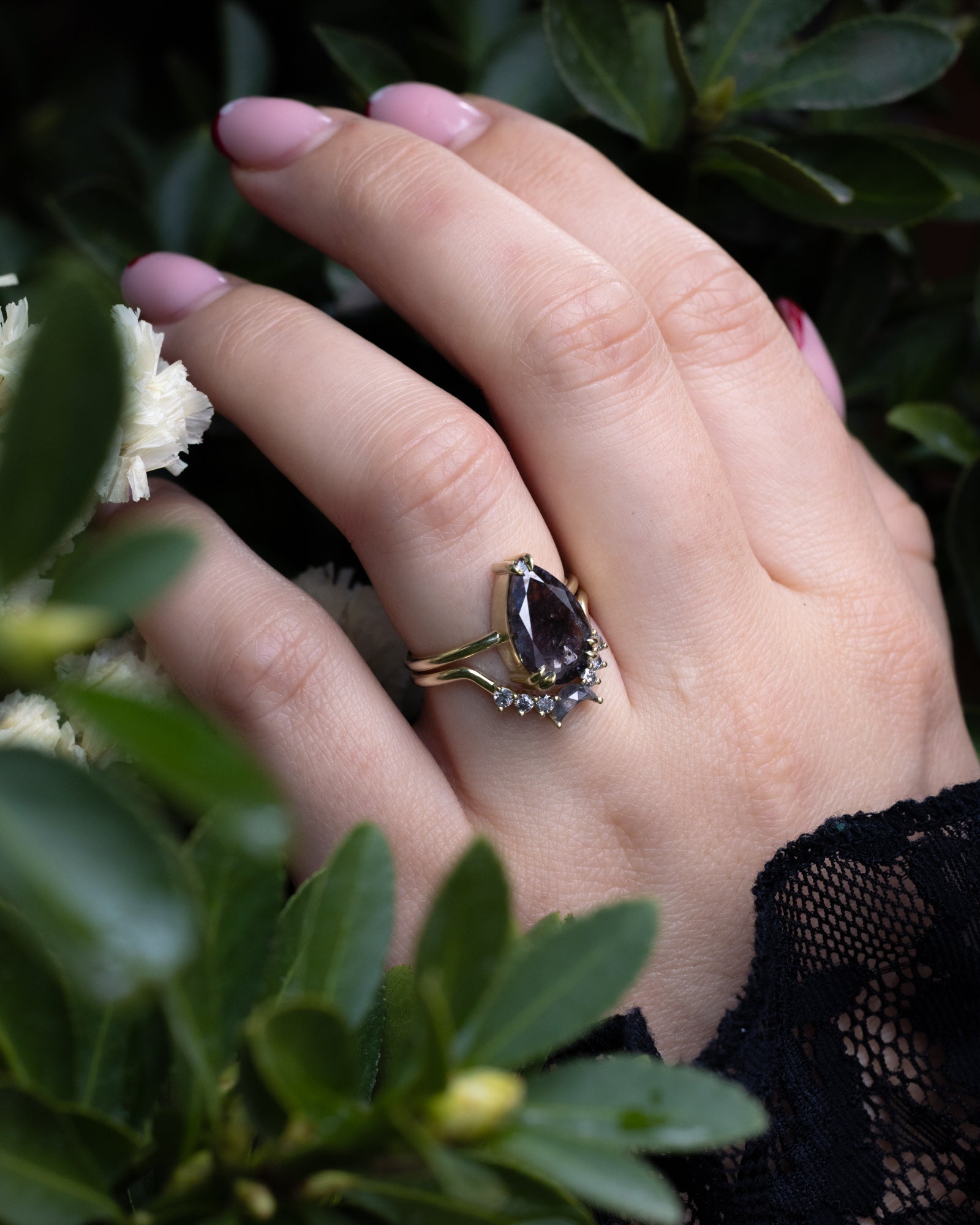 Hand wearing a ring with a salt & pepper diamond centerstone against a green leafy background