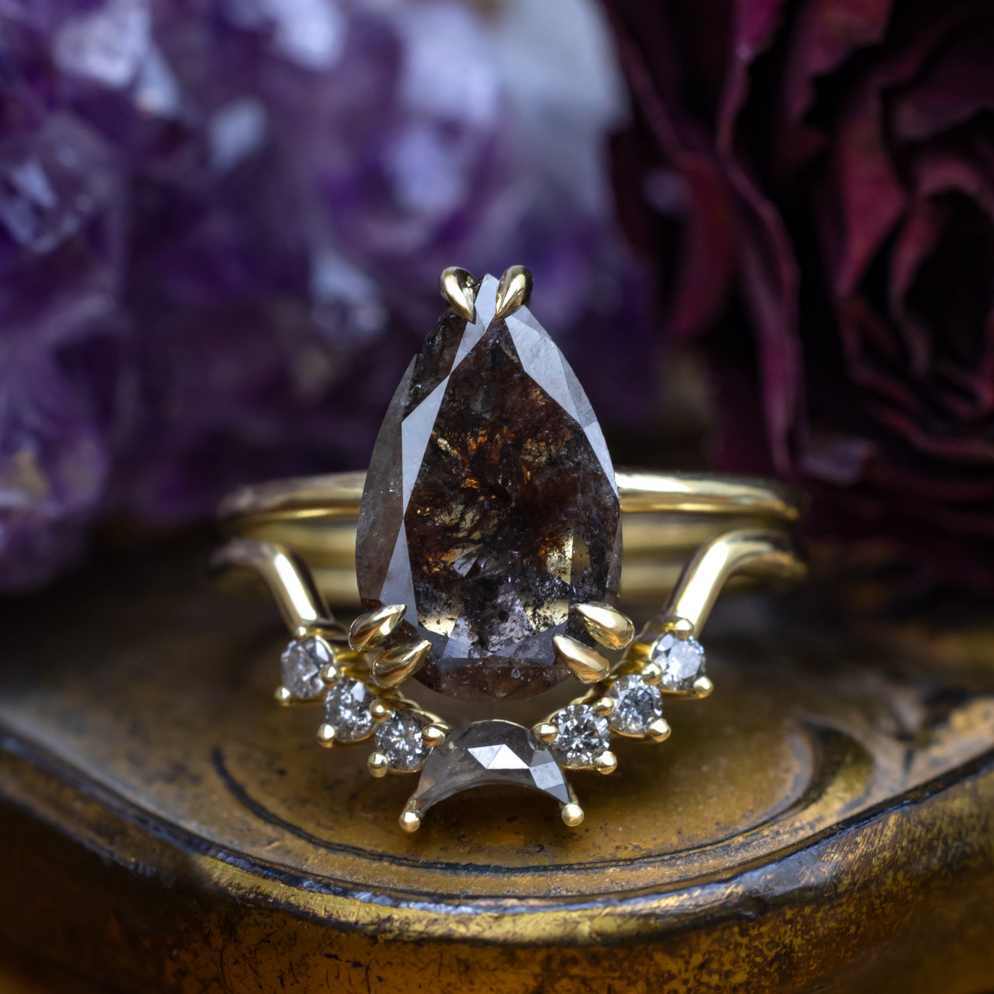 Green gold ring with a pear salt & pepper diamond centerstone and accents on a decorative stand with purple flowers in the background.