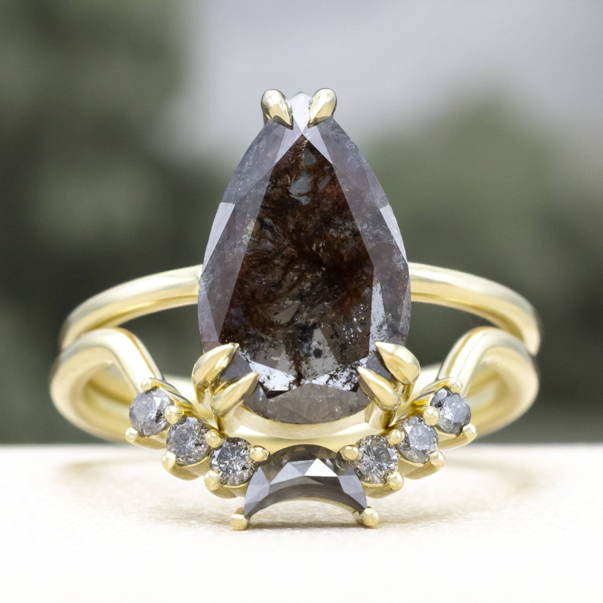 Green gold ring with a pear salt & pepper diamond centerstone and accents against a blurred background