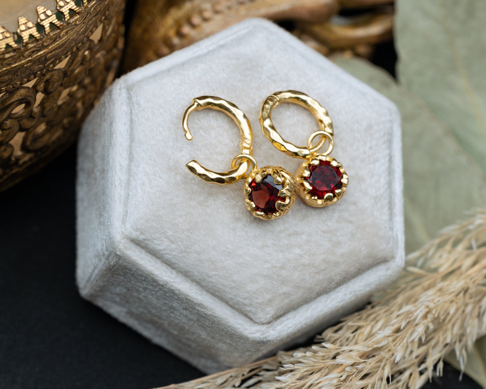 Gold hoop earrings with red garnets on a white hexagonal jewelry box.