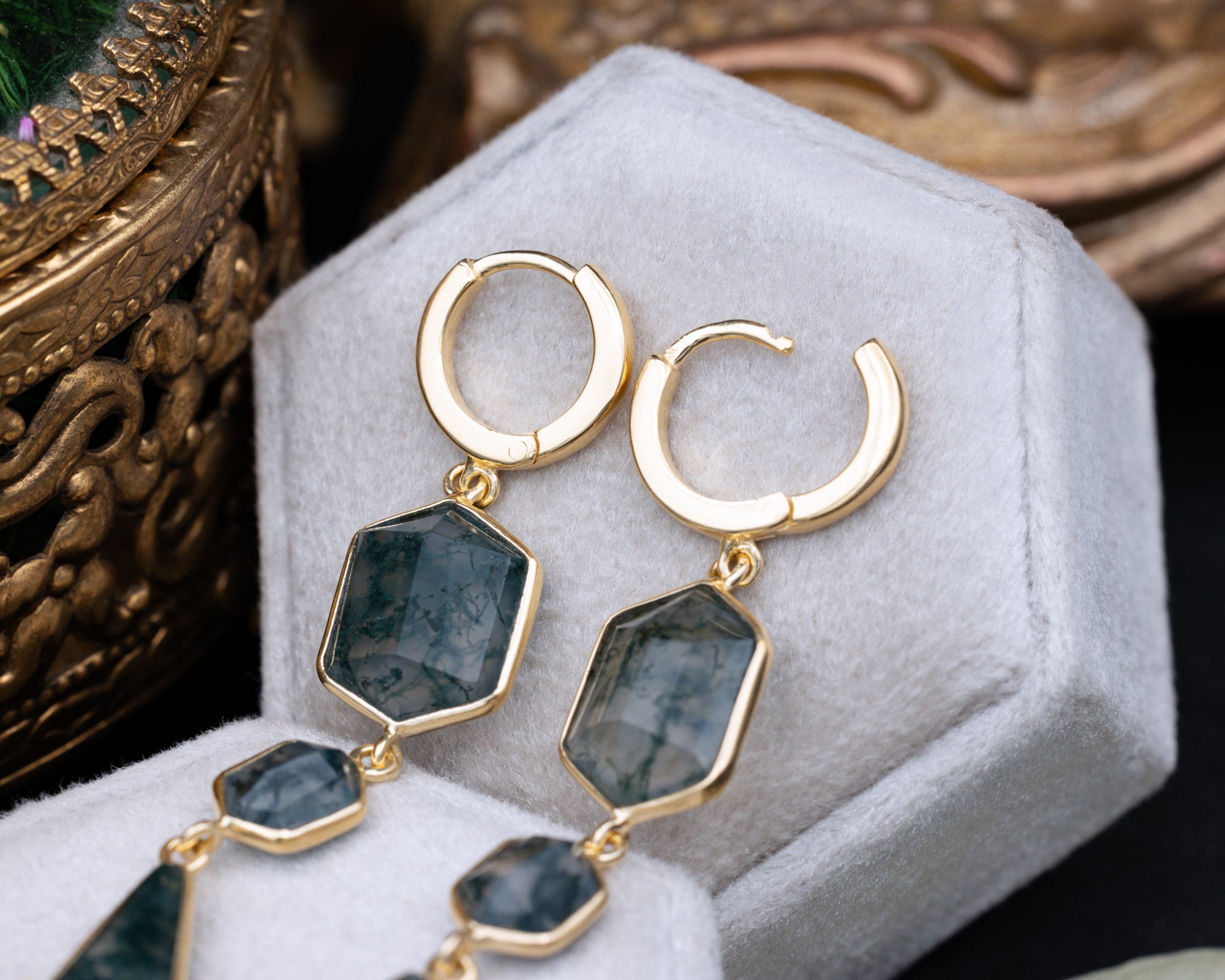 Gold moss agate earrings on a white jewelry box with decorative elements.