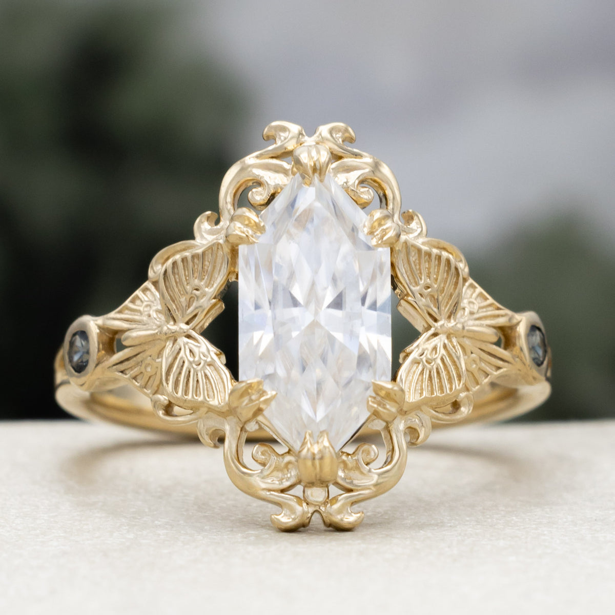 Gold ring with moissanite center stone and moth filigree against a blurred green background