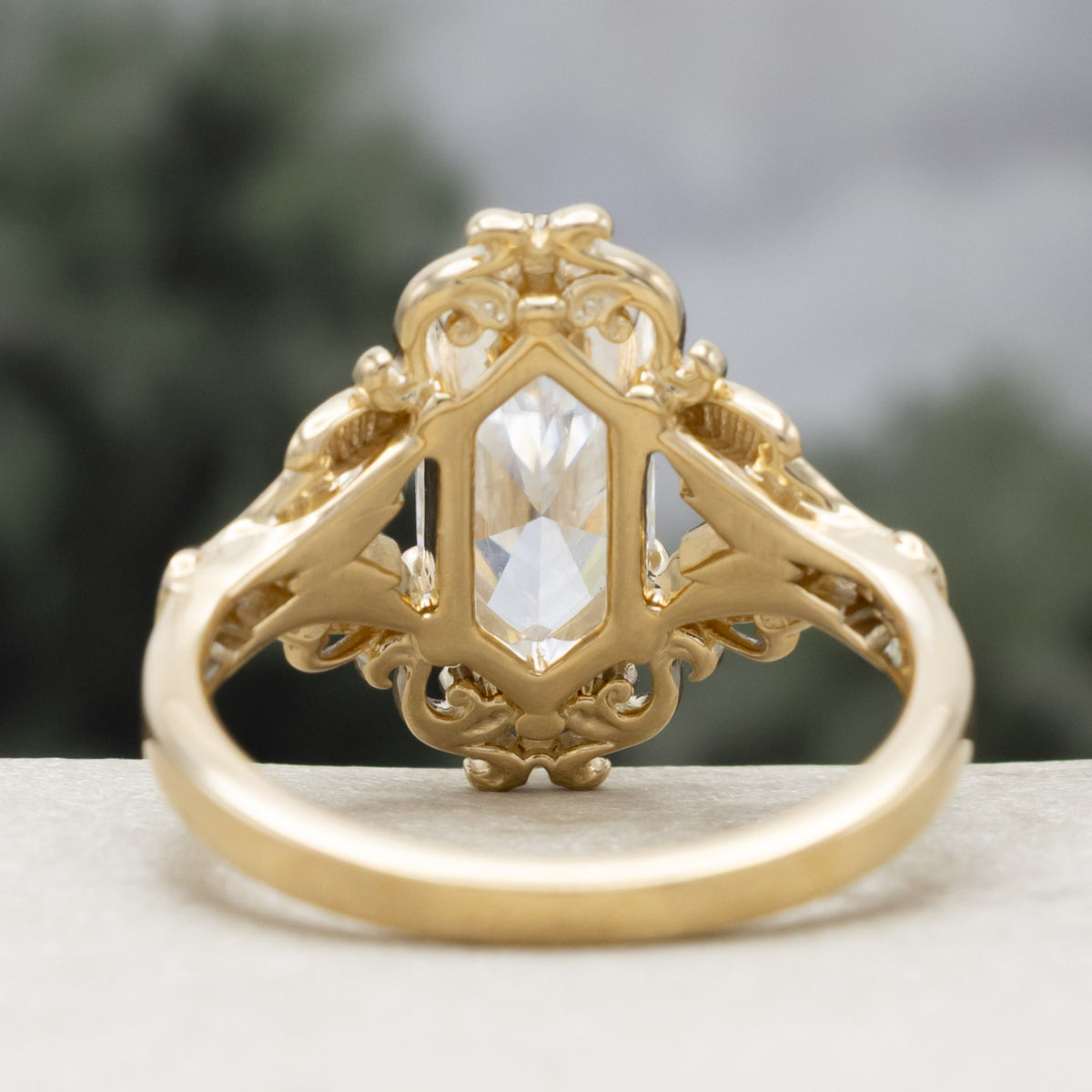 Gold ring with moissanite center stone and moth filigree against a blurred green background