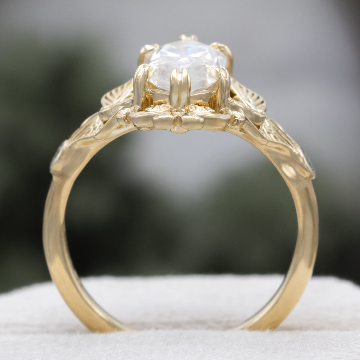 Gold ring with moissanite center stone and moth filigree against a blurred green background