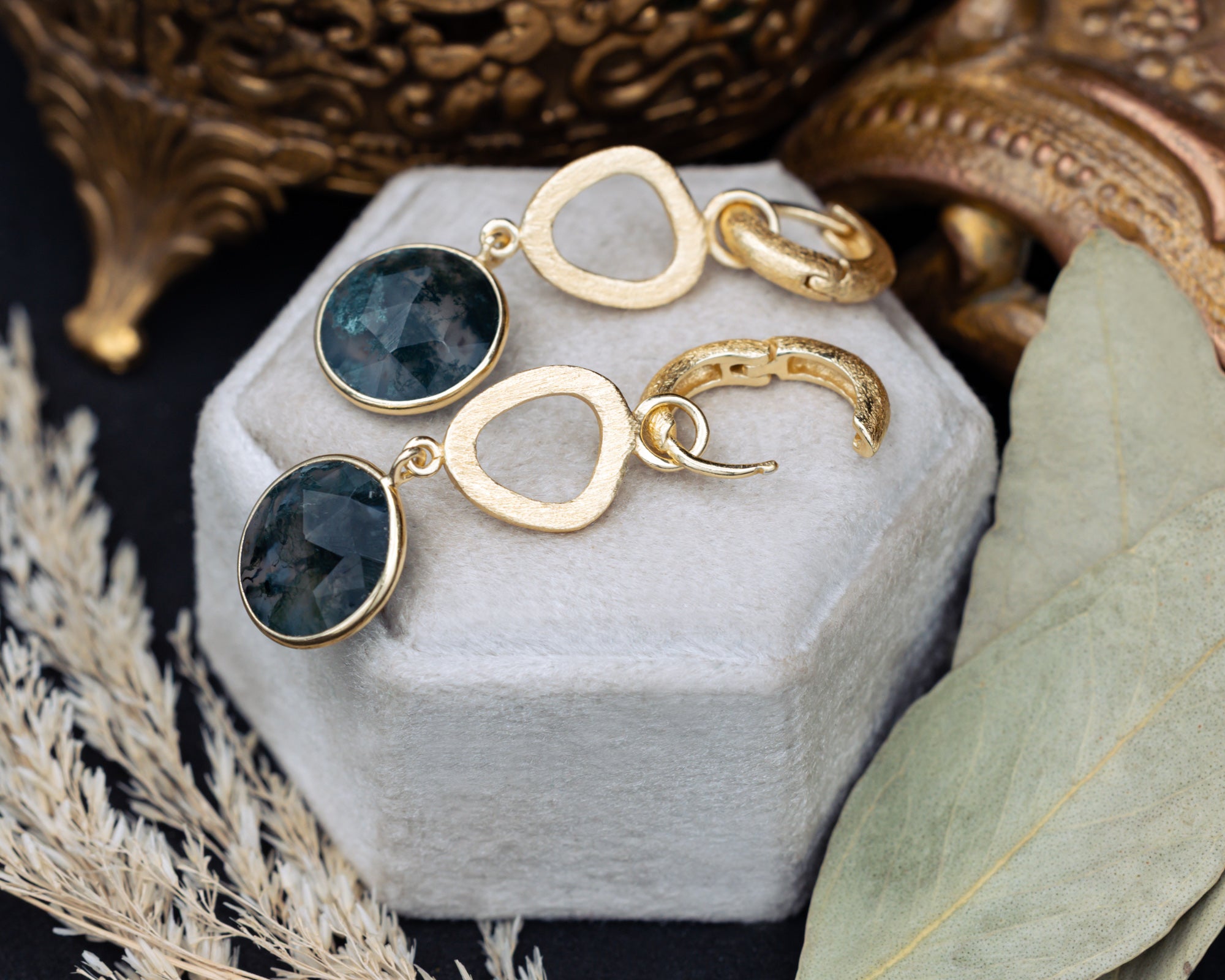 Gold earrings with moss agate gemstones on a white jewelry box with decorative background