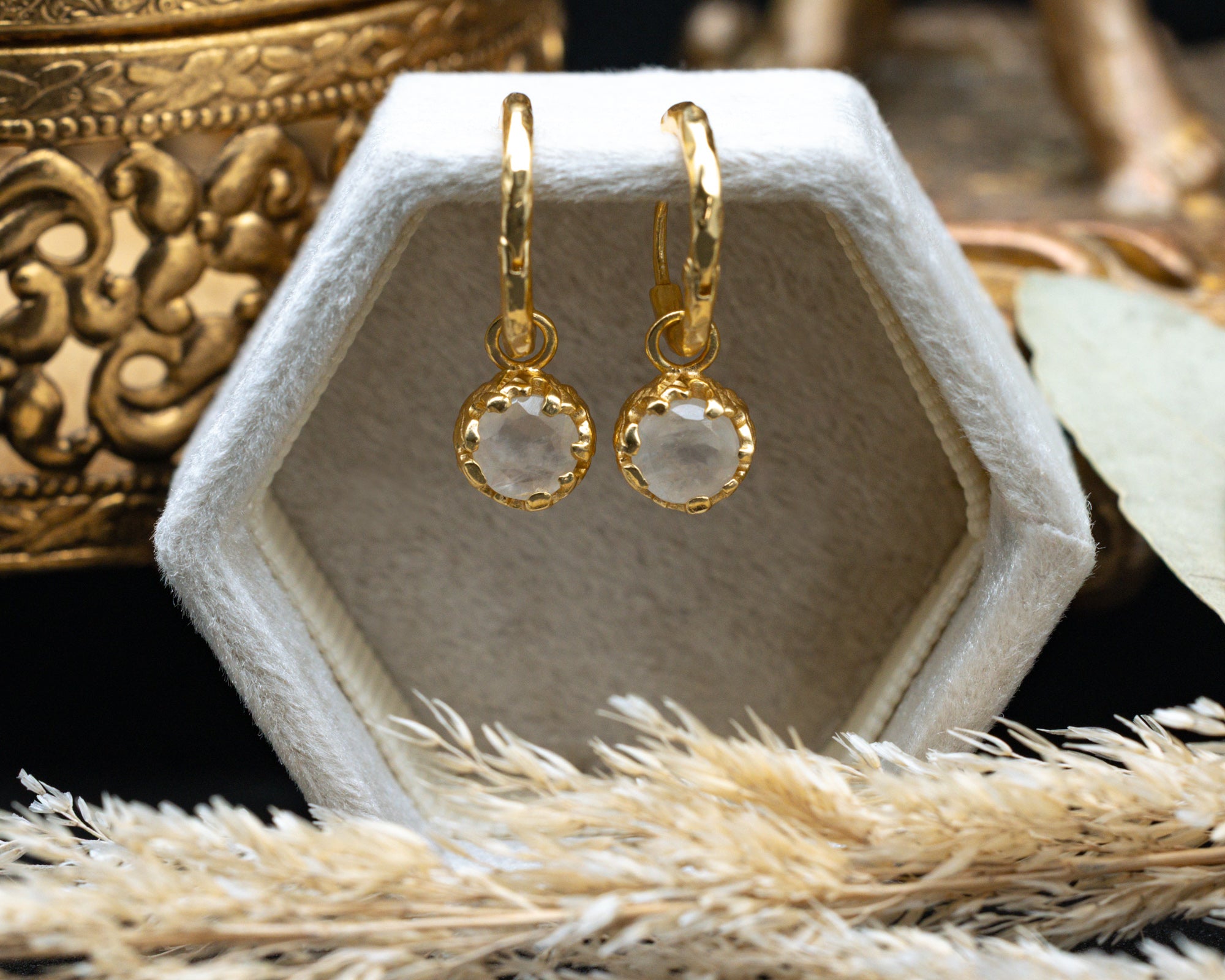 Gold earrings with moonstones on a hexagonal white stand, surrounded by decorative elements.