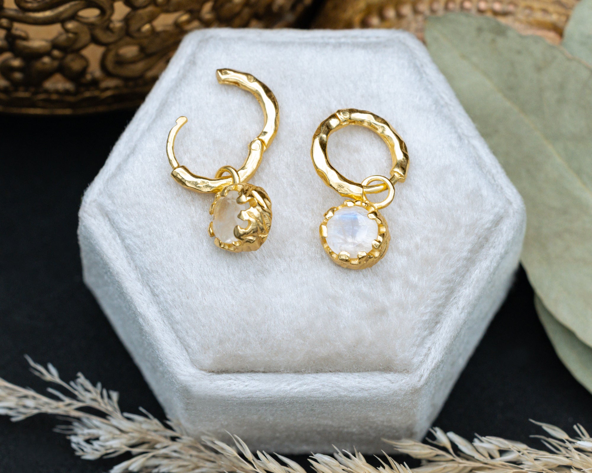Gold hoop earrings with a small moonstone on a white hexagonal jewelry box.