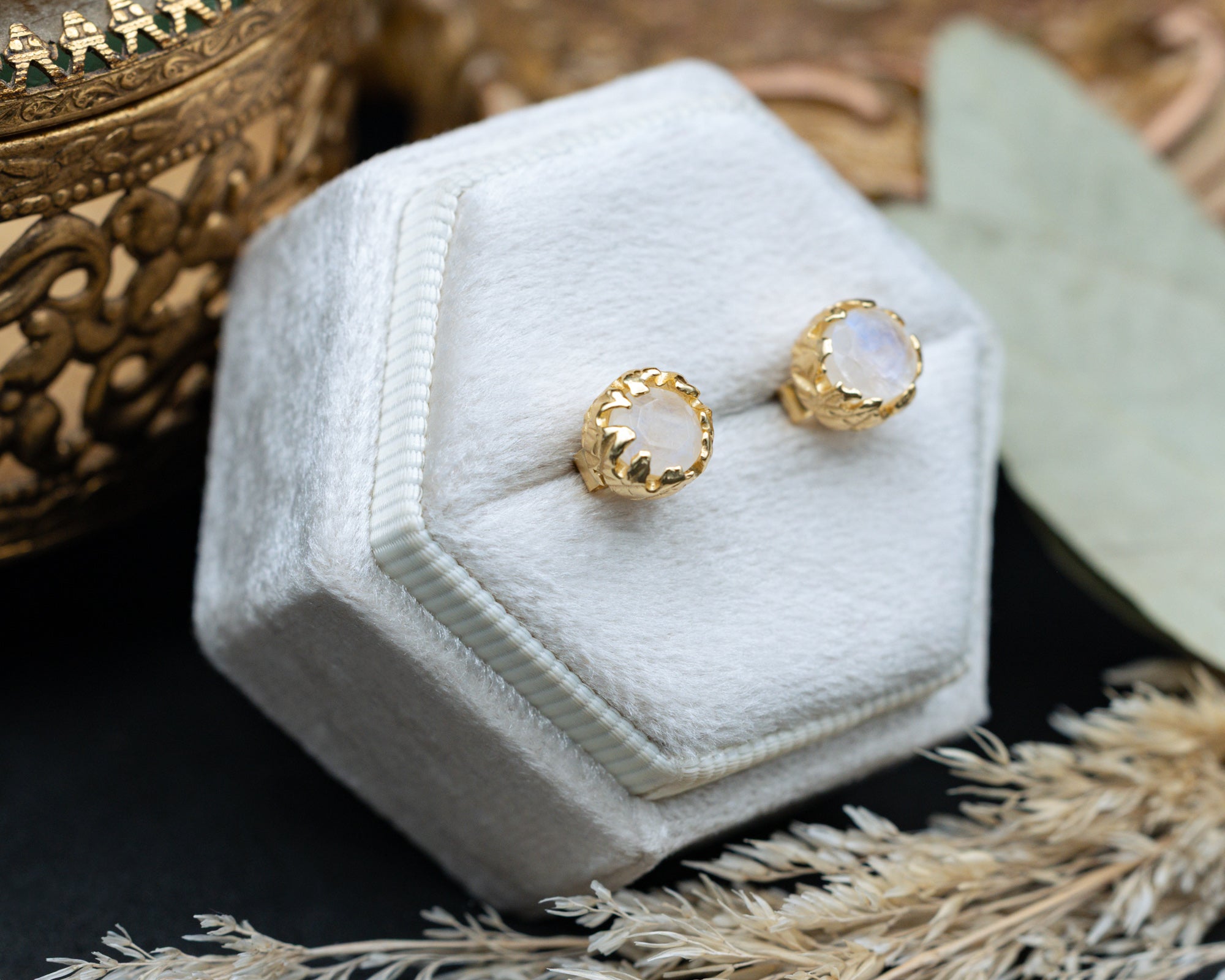 Gold moonstone earrings on a white hexagonal jewelry box with a decorative background.