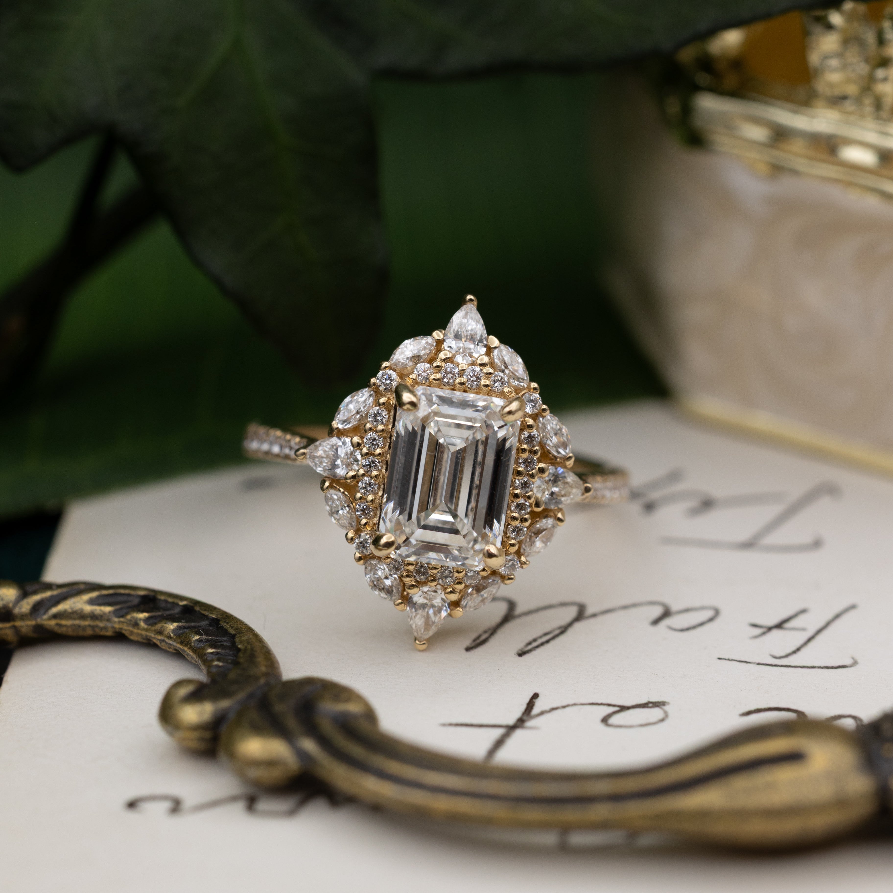 Diamond ring with emerald-cut center stone on a piece of paper with writing, surrounded by green leaves.