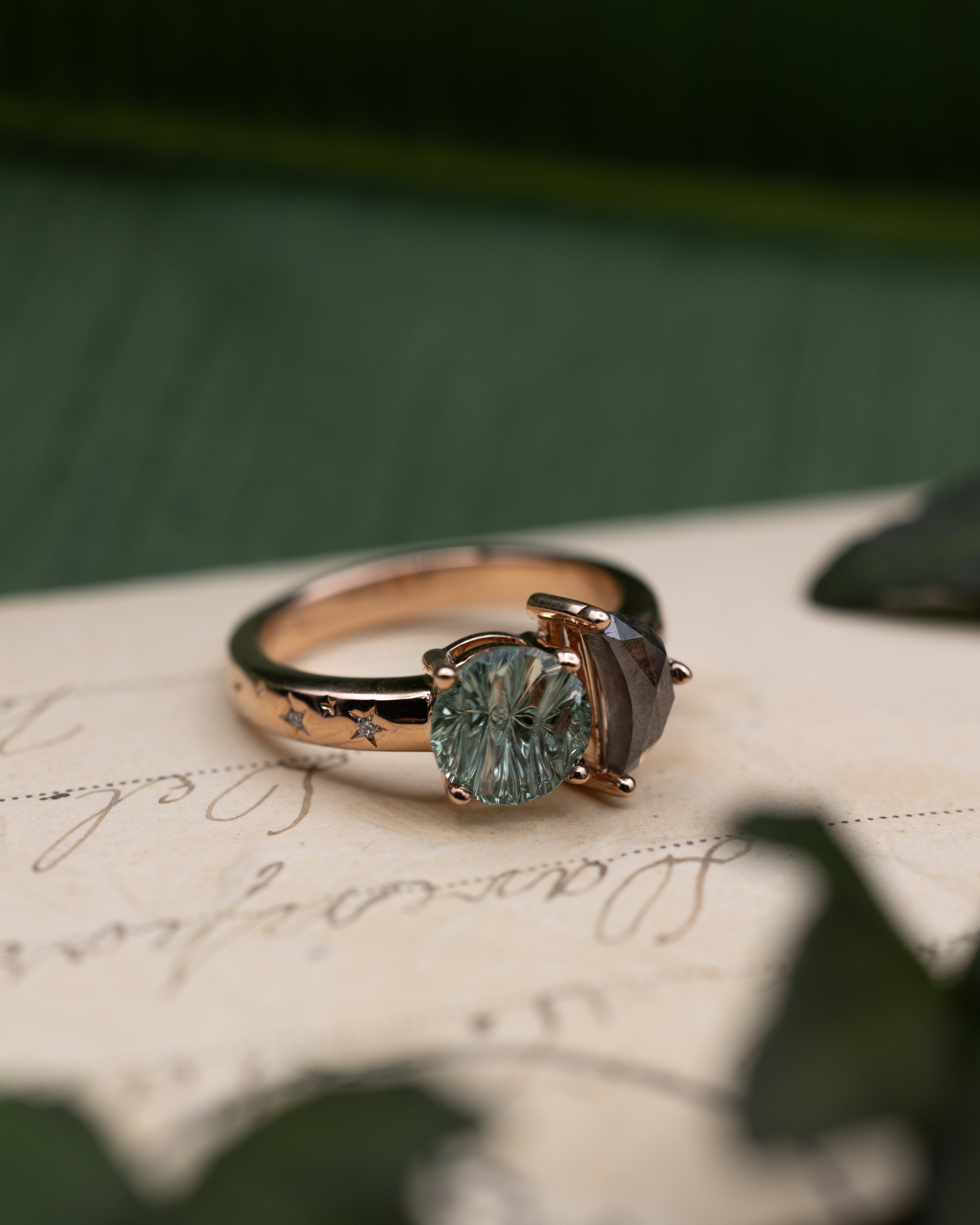 Rose gold ring with salt & pepper diamond crescent moon and round green sapphire on a vintage letter against a blurred green background