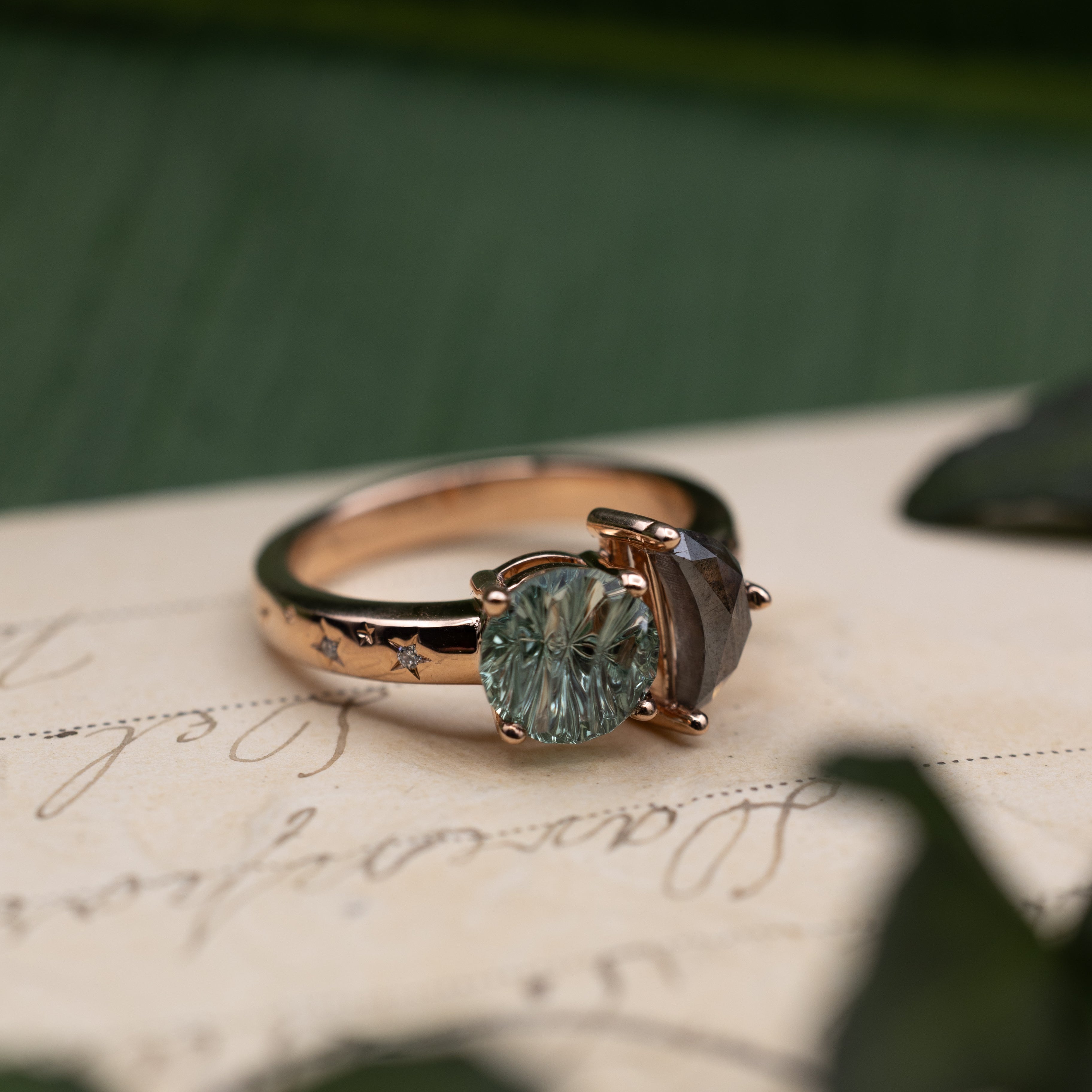 Rose gold ring with salt & pepper diamond crescent moon and round green sapphire on a vintage letter against a blurred green background