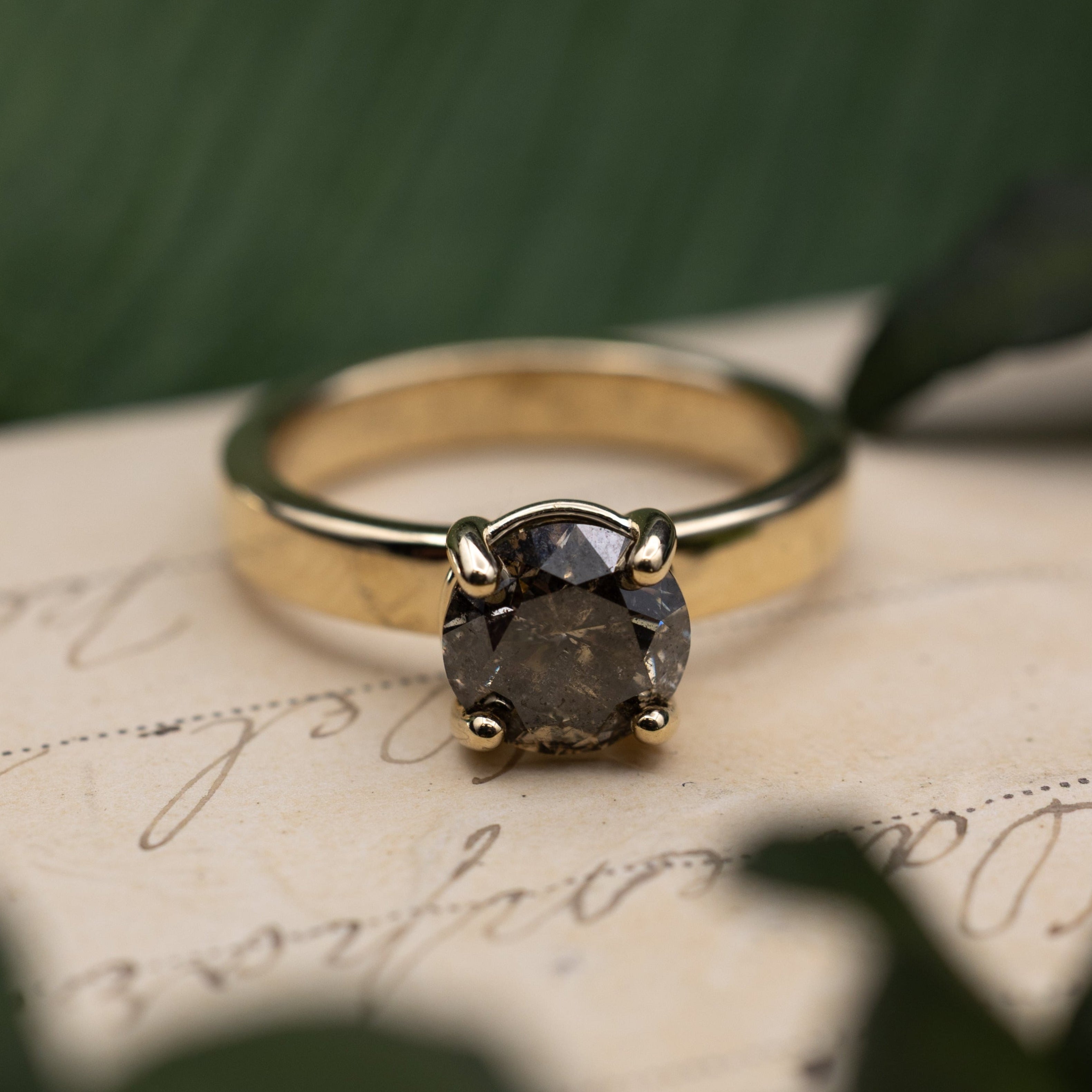 Yellow gold ring with salt & pepper diamond center stone sitting on a vintage letter against a blurred green background