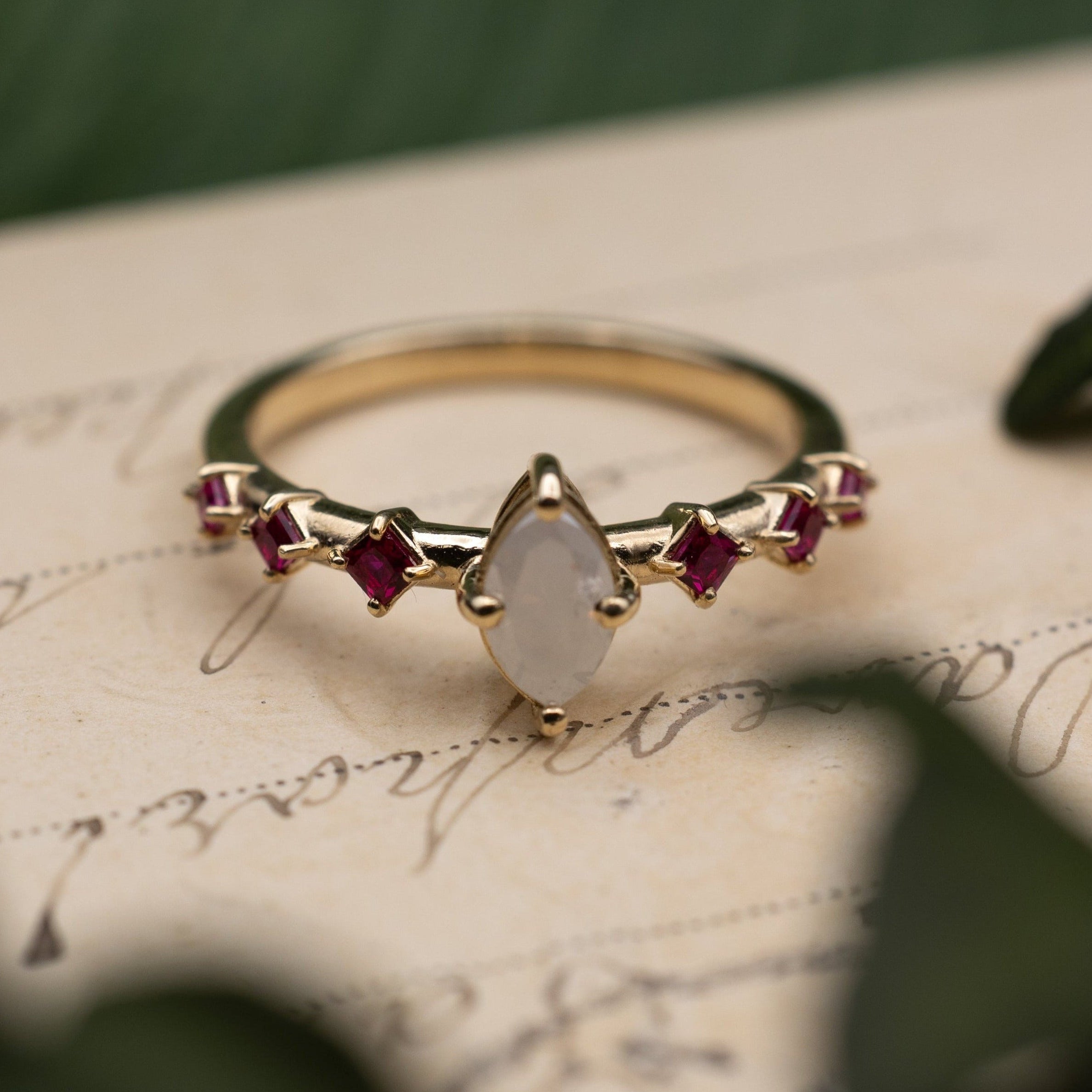 Yellow gold ring with salt & pepper diamond center stone and ruby accents on a vintage letter against a blurred green background