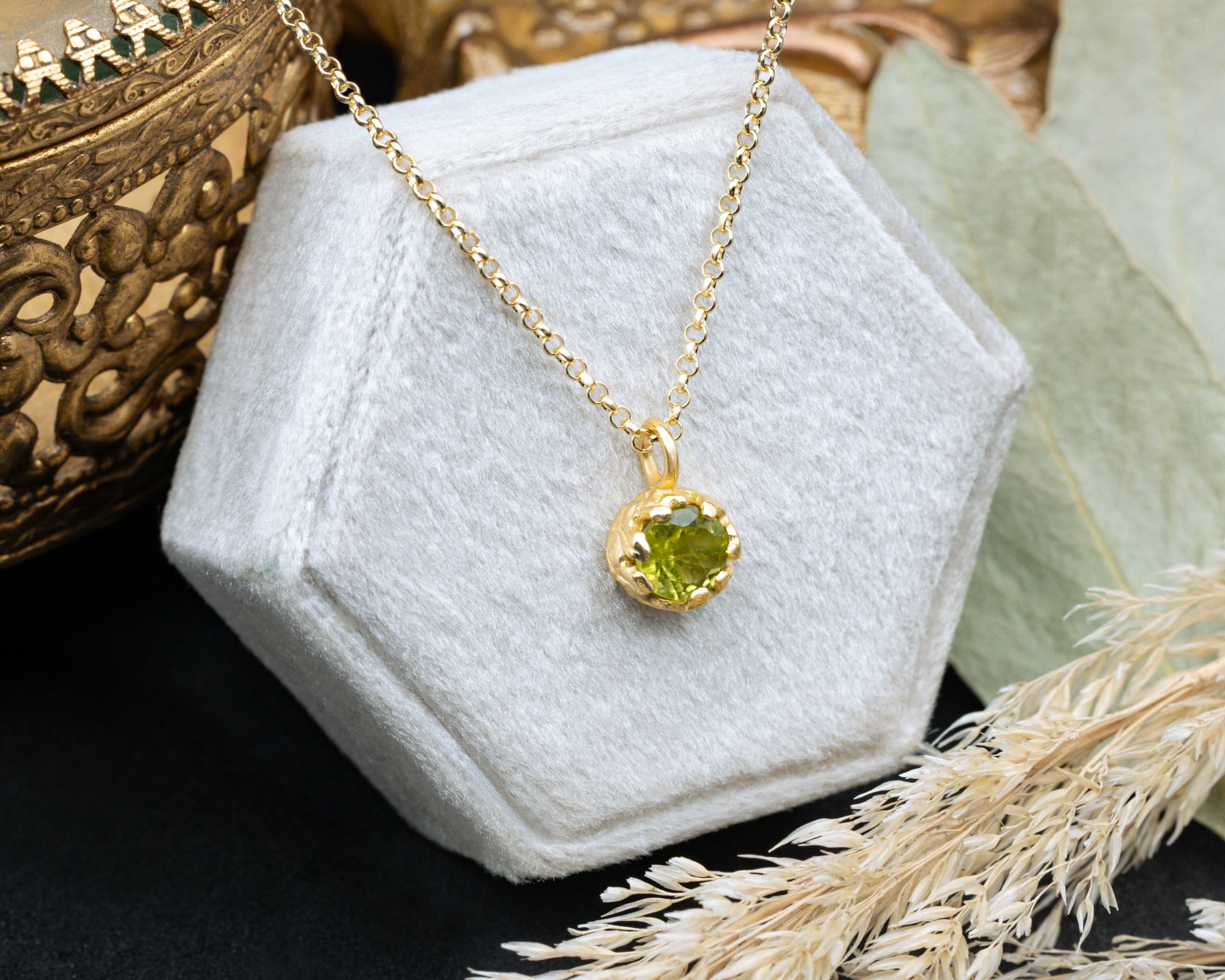 Gold necklace with a green peridot on a white hexagonal stand, with a decorative background.