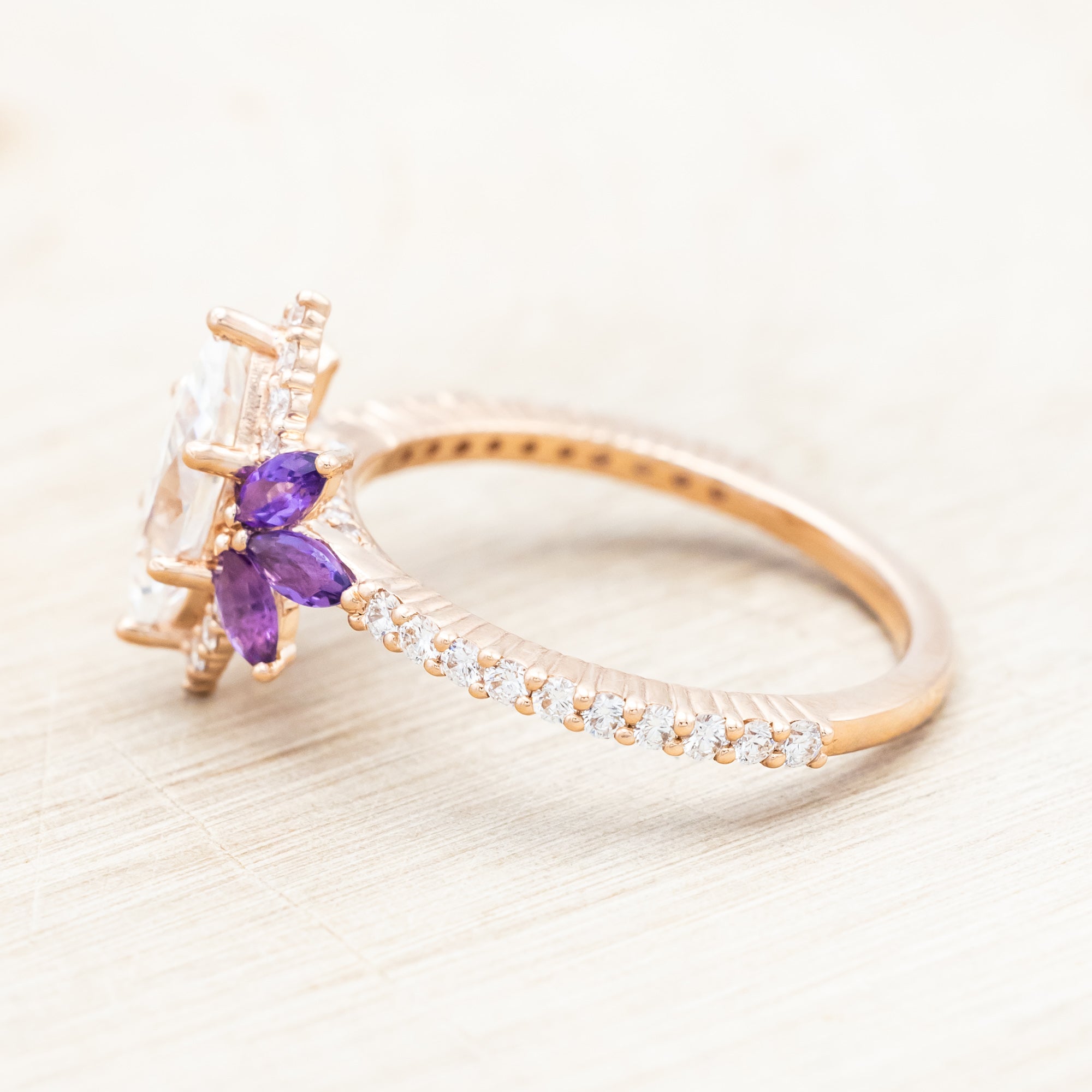 14K Rose gold ring with a moissanite center stone and small purple amethyst accents on a light wooden background