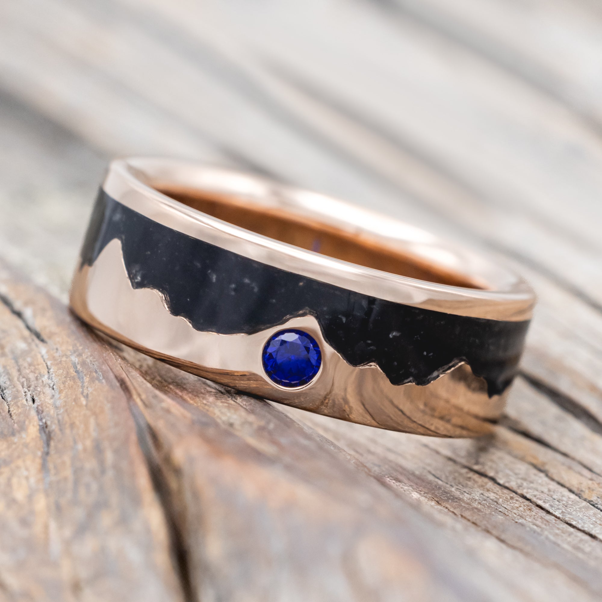 Rose gold ring with black inlay and blue sapphire on a wooden surface