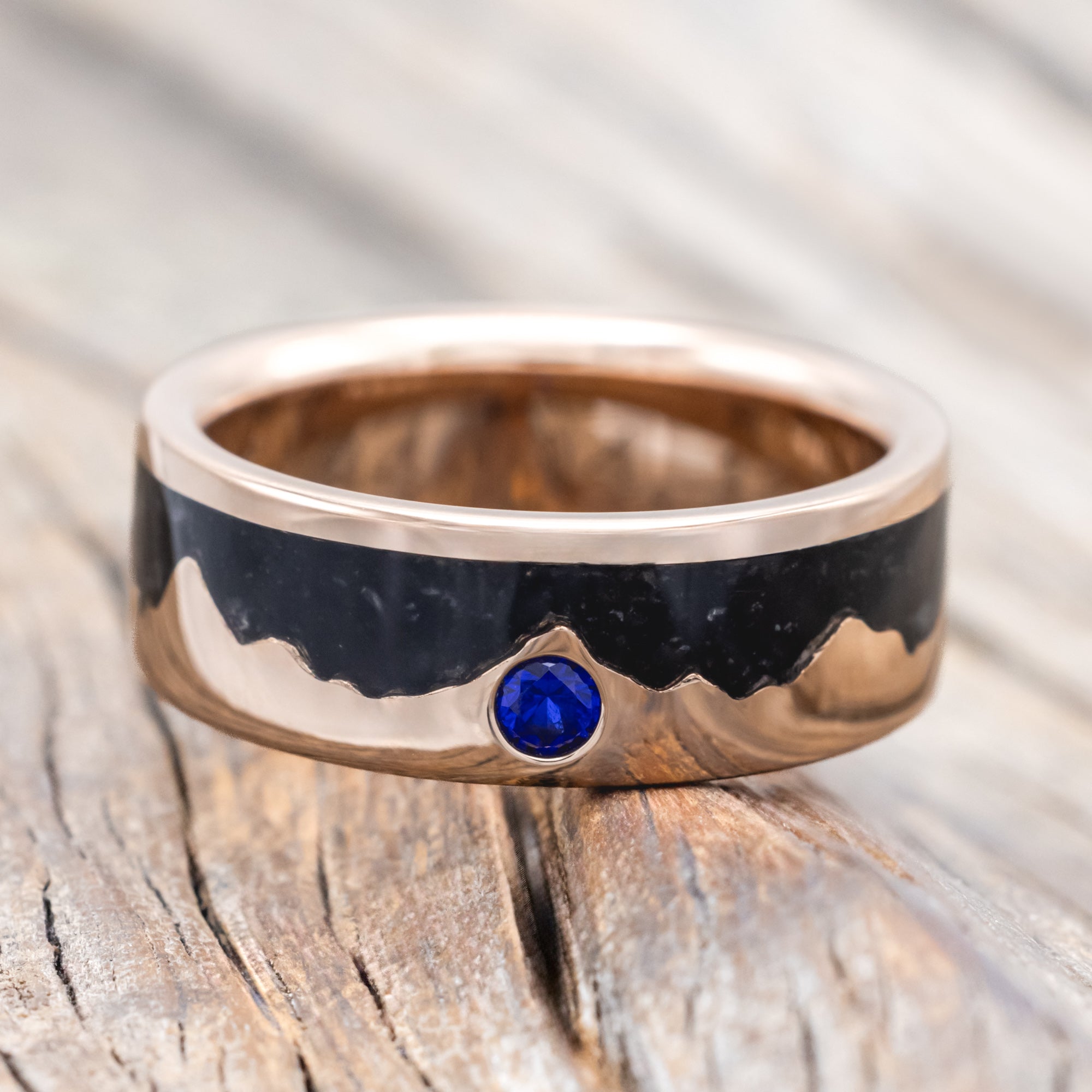 Rose gold ring with black inlay and blue sapphire on a wooden surface