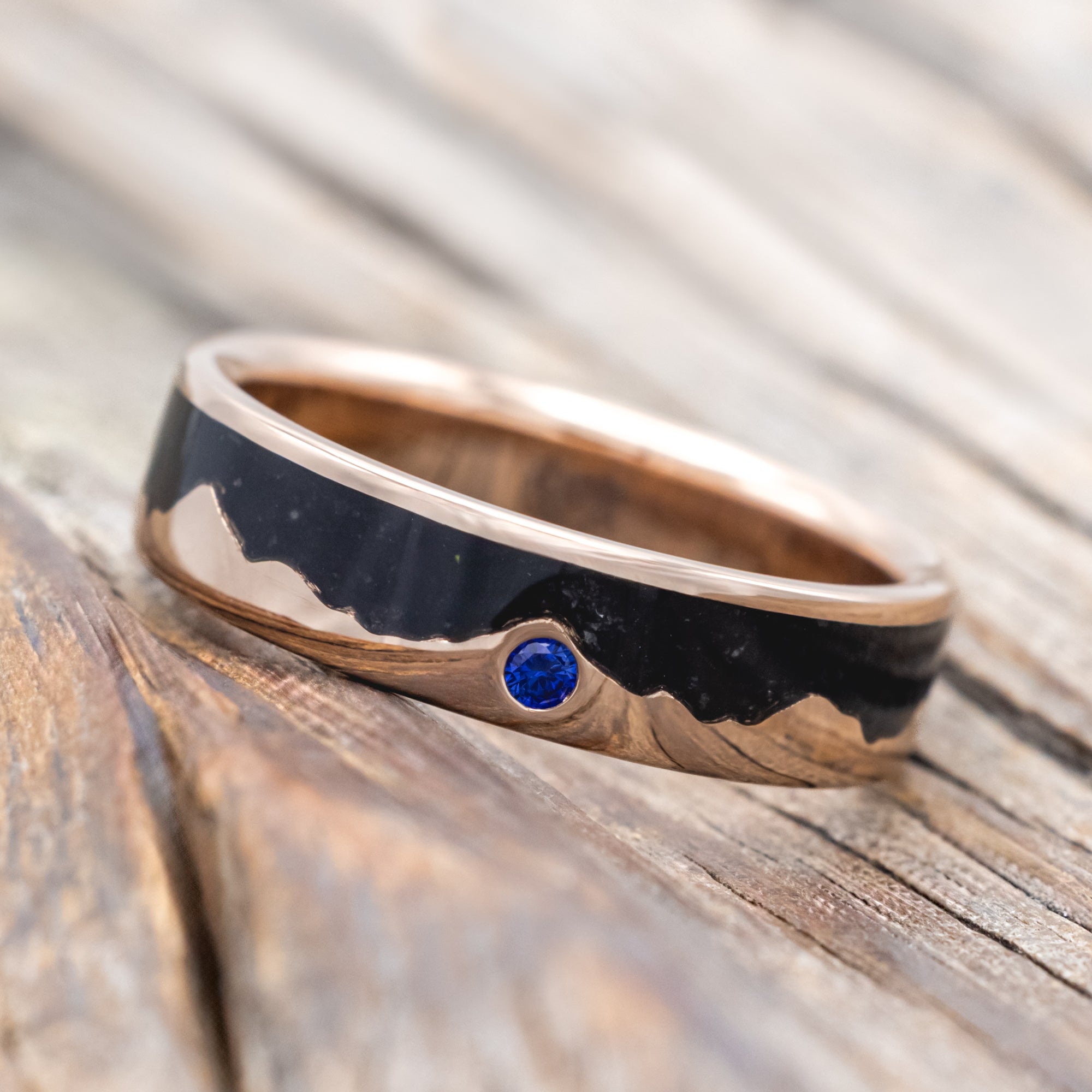 Rose gold ring with black inlay and blue sapphire on a wooden surface