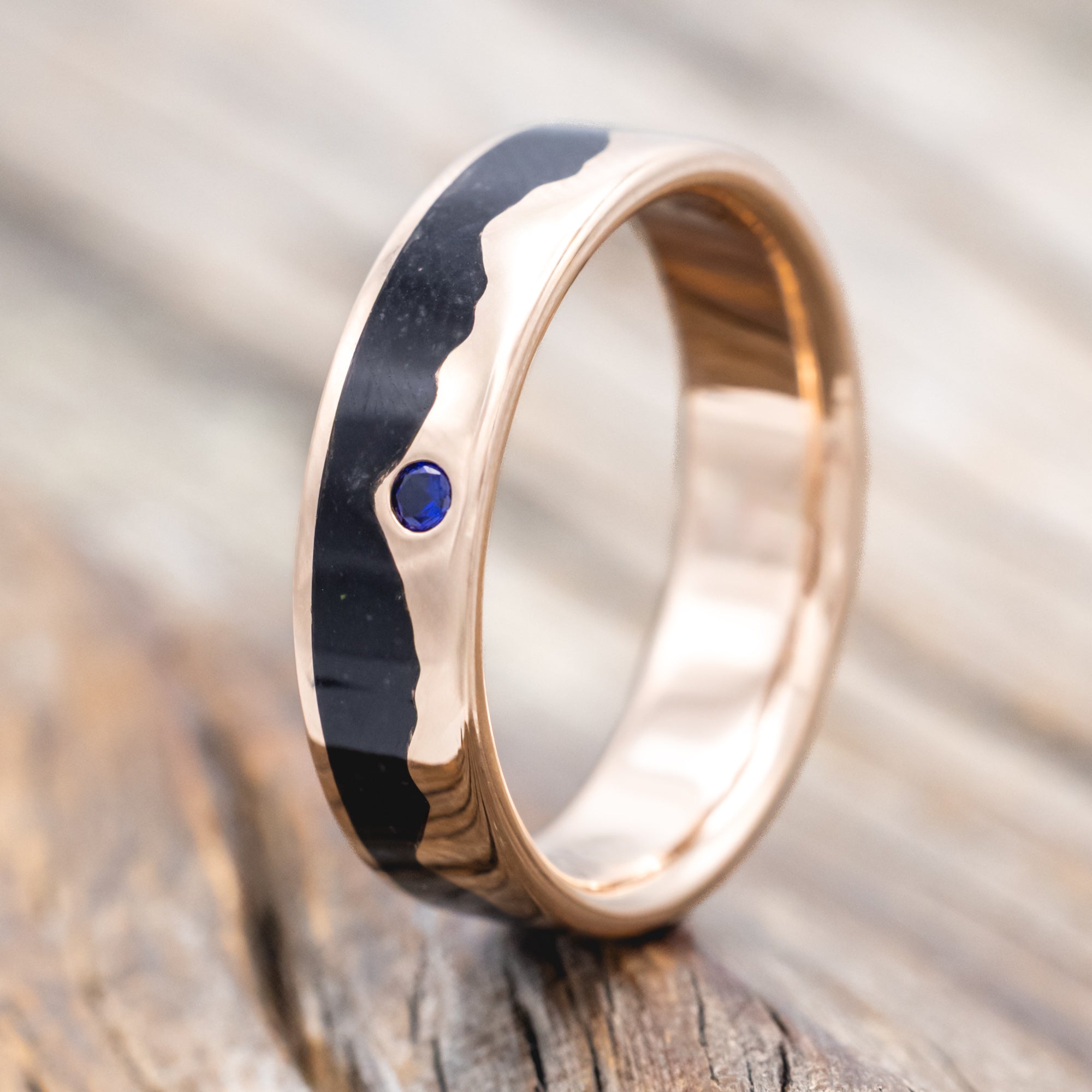 Rose gold ring with black inlay and blue sapphire on a wooden surface