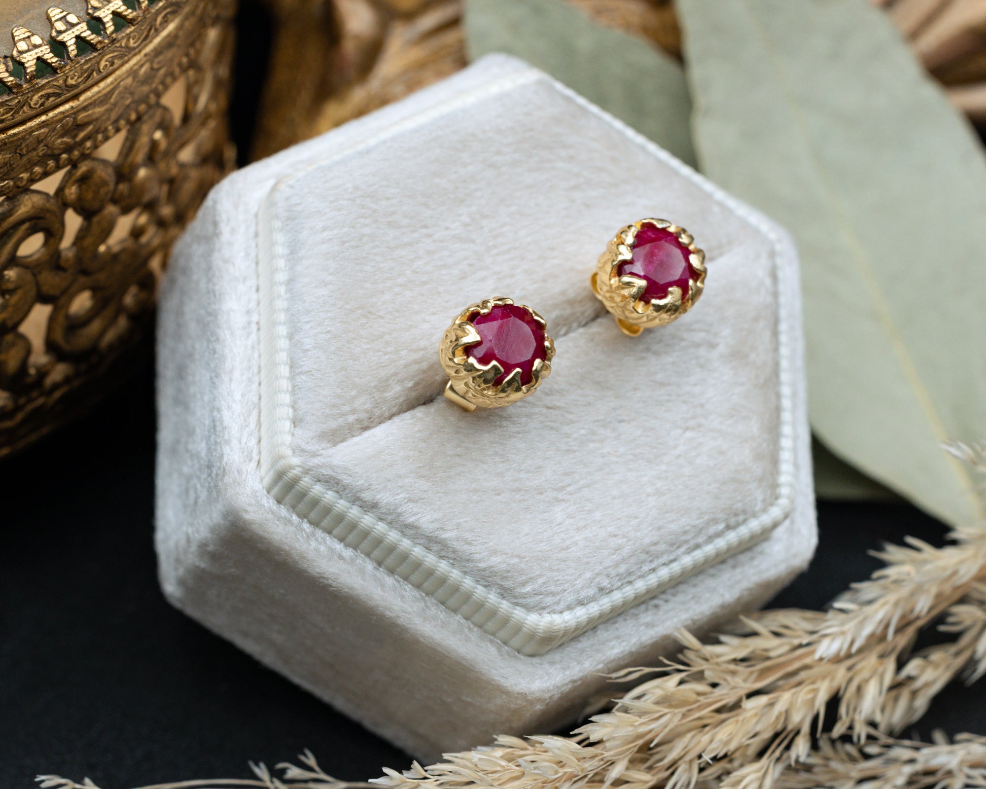 Gold earrings with red rubies in a white jewelry box on a dark surface.