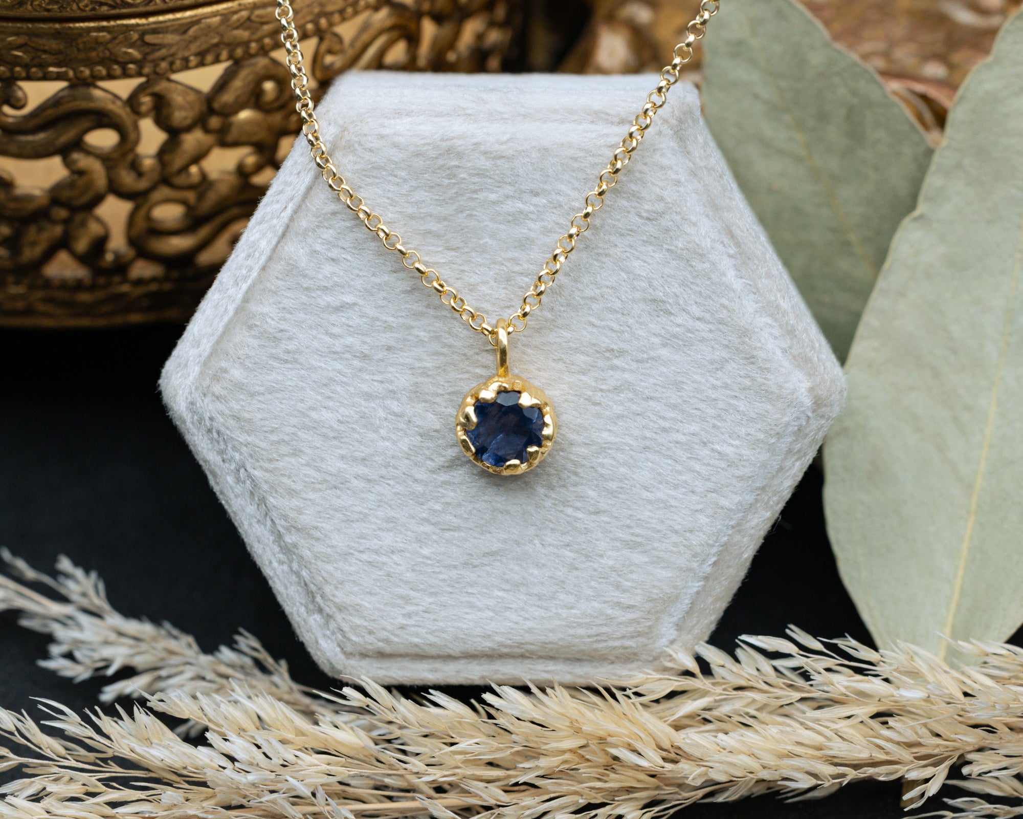 Gold necklace with a blue sapphire pendant on a white hexagonal stand, surrounded by decorative elements.