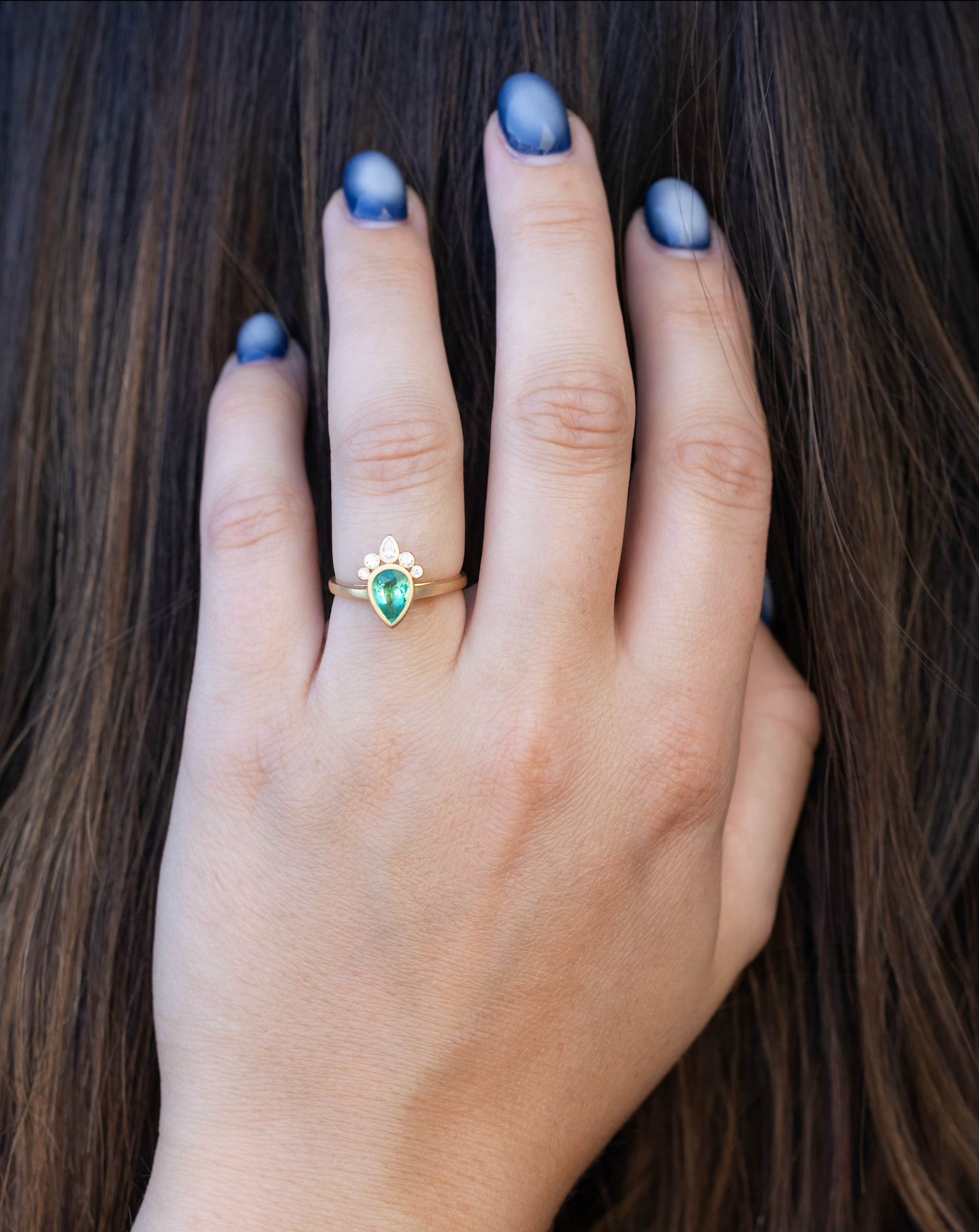Gold ring with emerald center stone and diamond accents on hand against dark hair