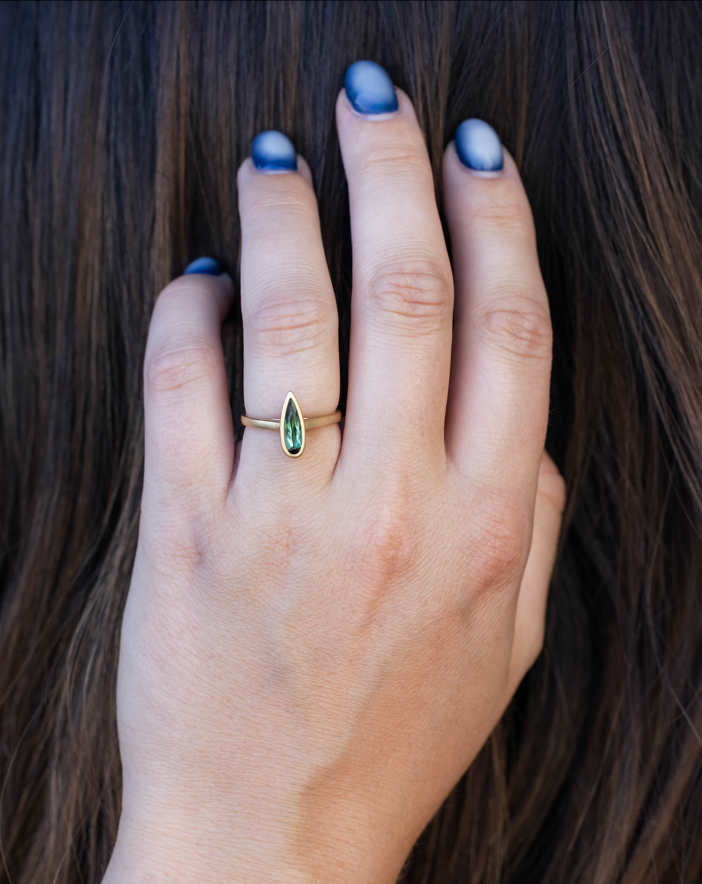 Hand wearing a gold ring with a green gemstone, set against dark hair.