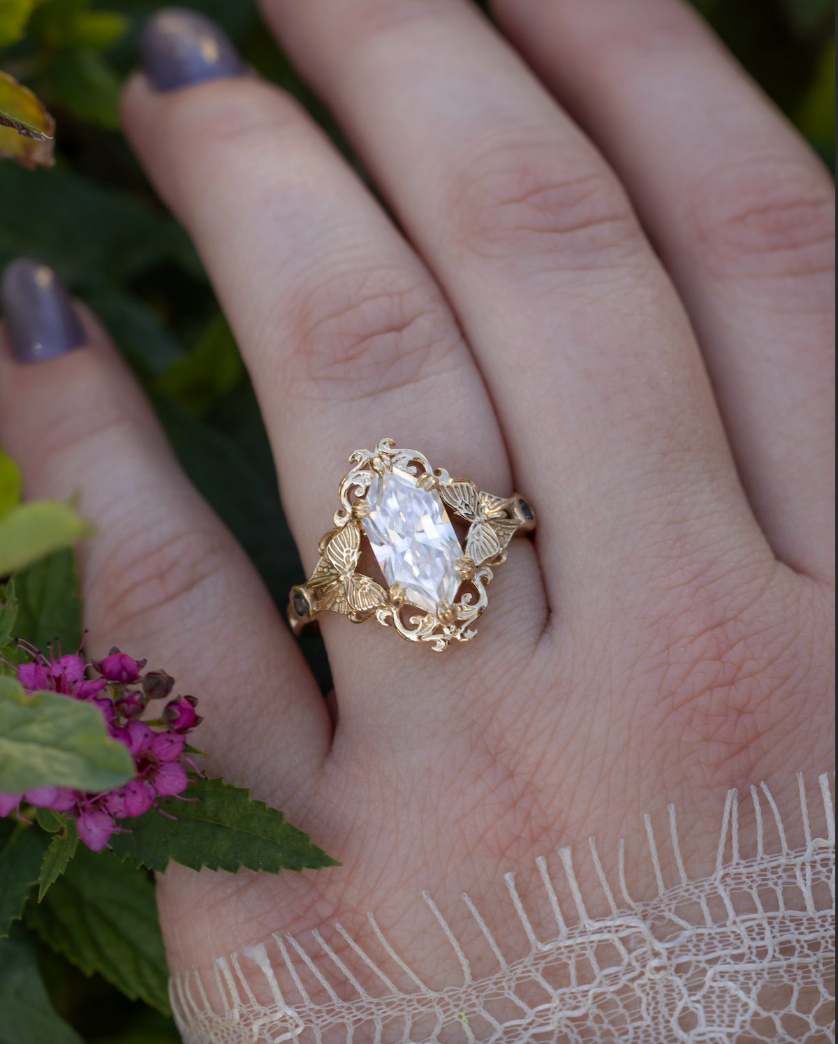 Hand wearing a gold ring with moissanite center stone, moth filigree and sapphire accents against a foliage background