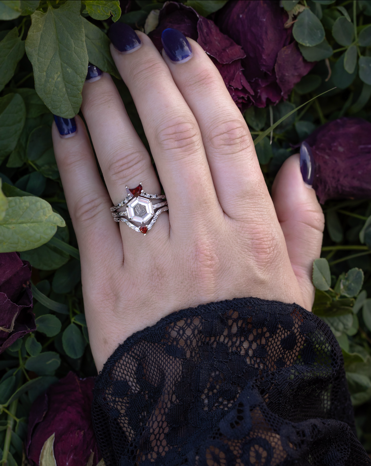 Hand wearing white gold engagement ring with portrait cut moissanite featuring rose design and matching garnet tracers on vine band against a foliage background