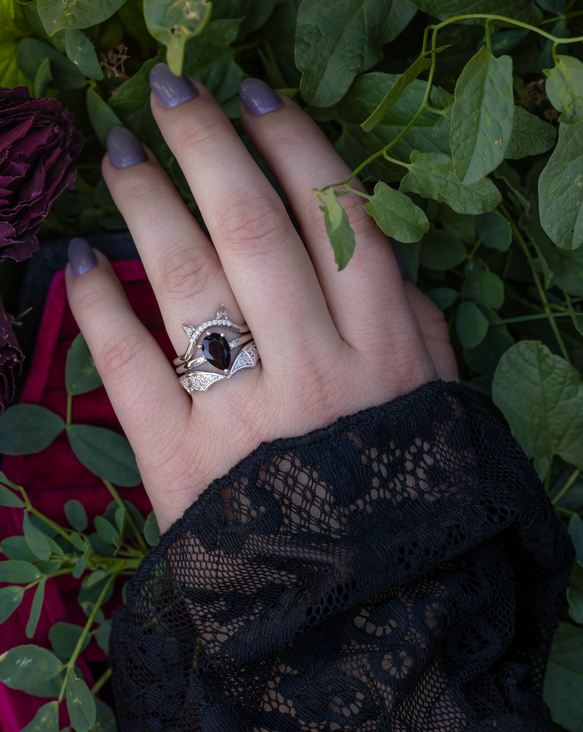Hand with black lace sleeve wearing a bat-inspired white gold bridal suite with smoky quartz center stone against a leafy background