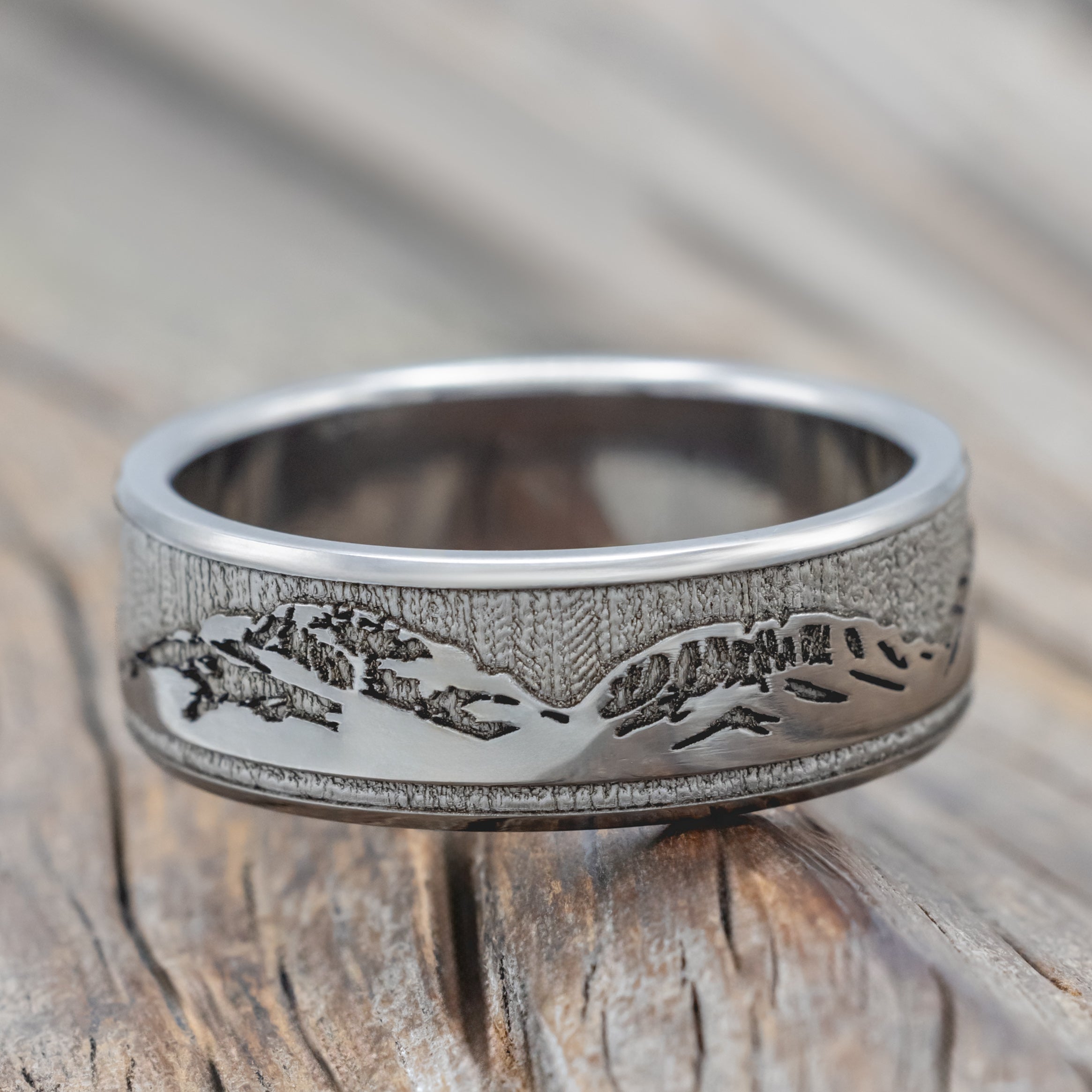 A titanium mountain ring with an engraved landscape design, resting on a rustic wooden surface.