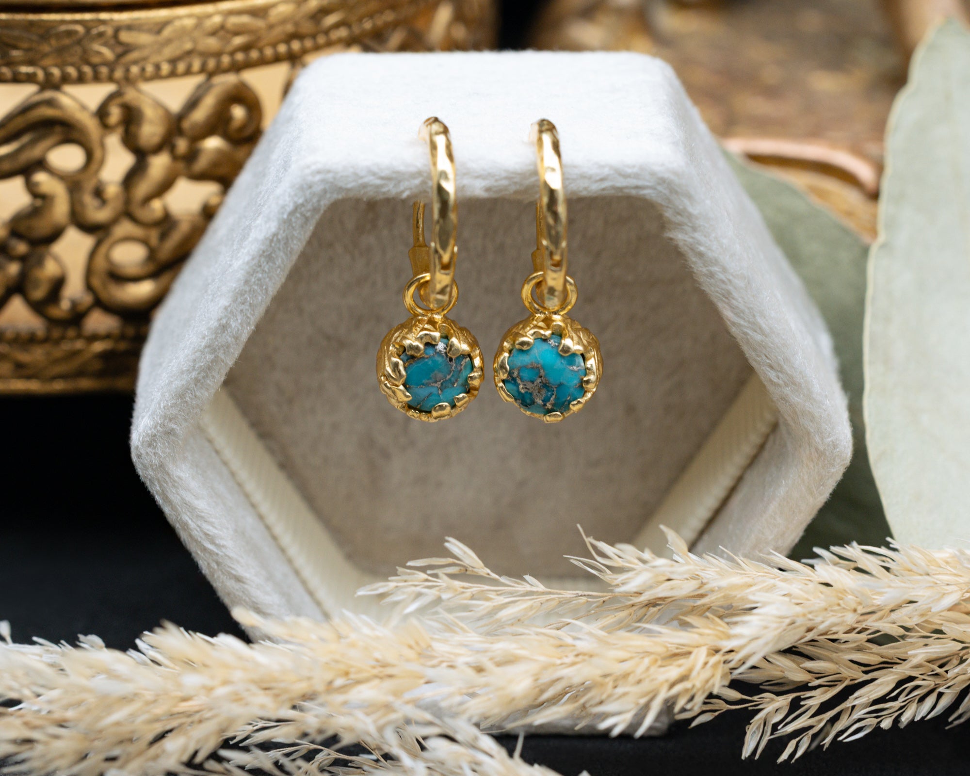 Gold earrings with turquoise stones on a white hexagonal stand.