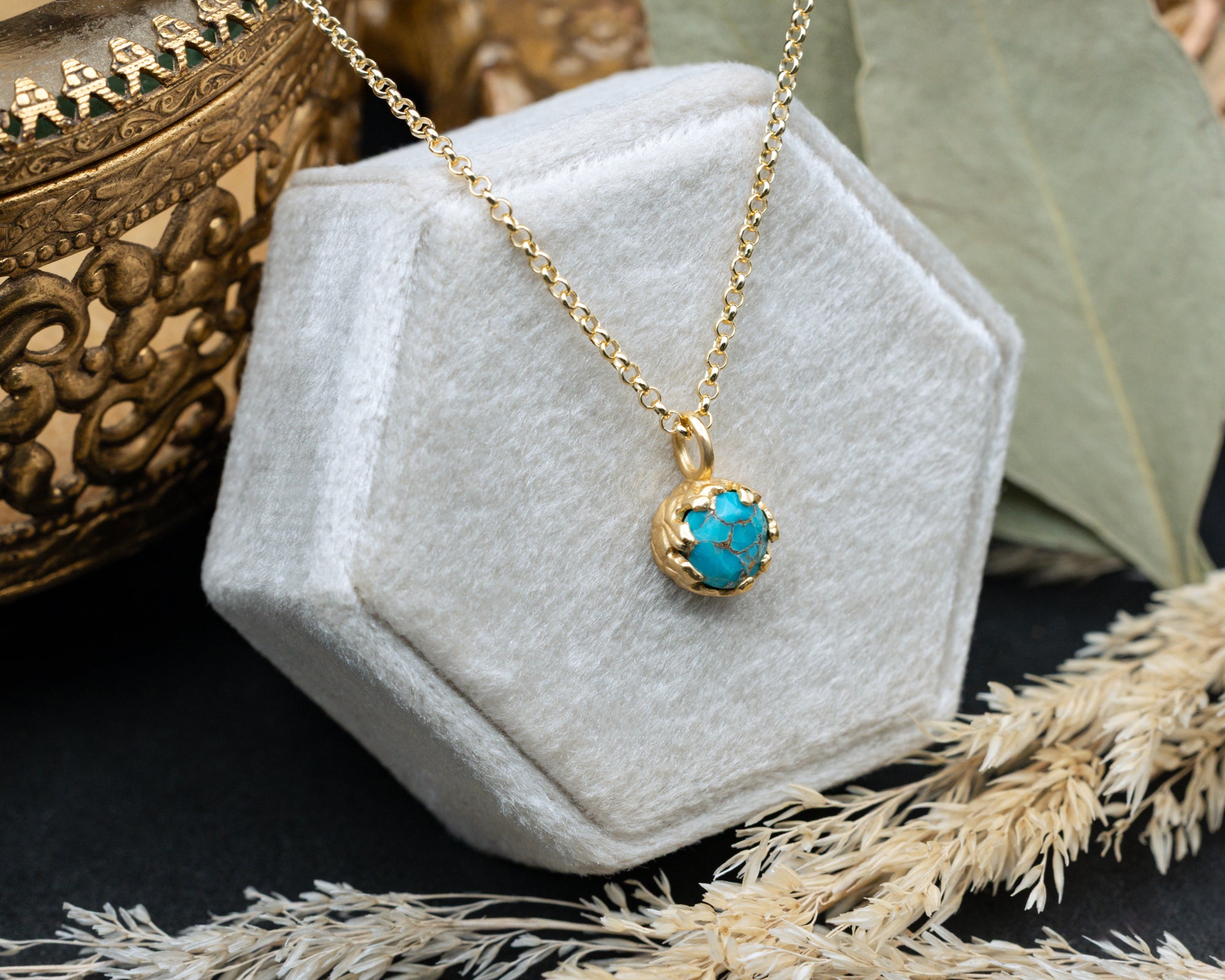 Gold necklace with a turquoise pendant on a white hexagonal stand, surrounded by decorative elements.