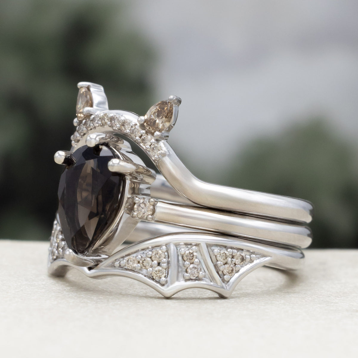 White gold engagement ring with smoky quartz center stone, bat ear top tracer, and bat wing bottom tracer with champagne diamond accents against a blurred green background