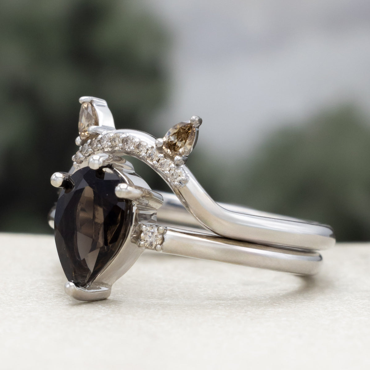White gold engagement ring with smoky quartz center stone and bat ear top tracer with champagne diamond accents against a blurred green background