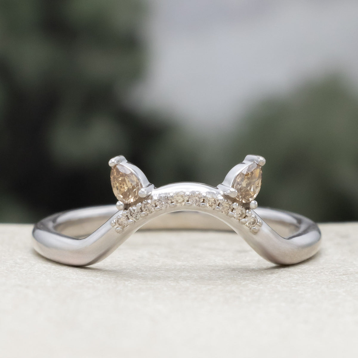 White gold cat ear wedding ring tracer with champagne diamond accents against a blurred green background