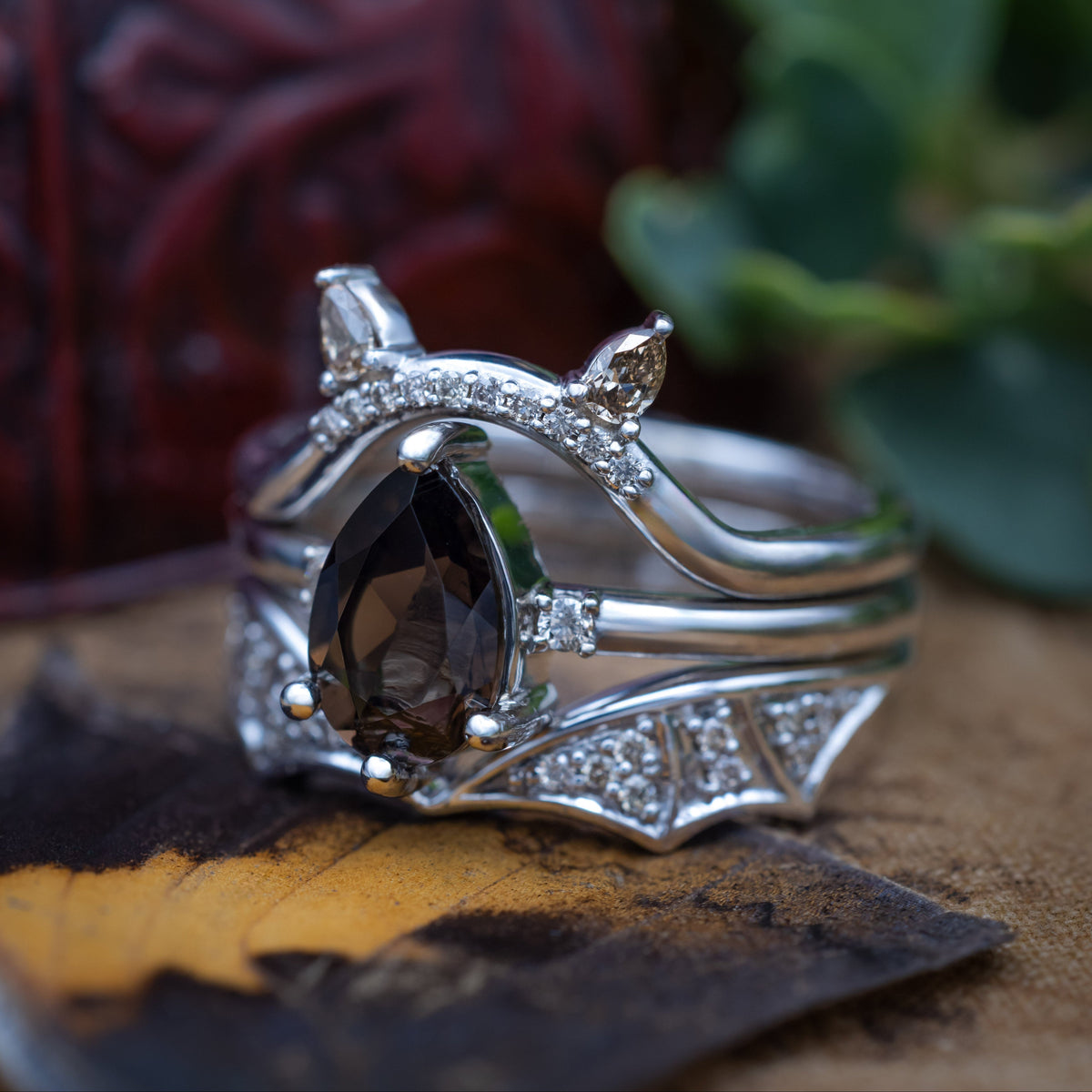 White gold engagement ring with smoky quartz center stone, bat ear top tracer, and bat wing bottom tracer with champagne diamond accents against a decorative green background