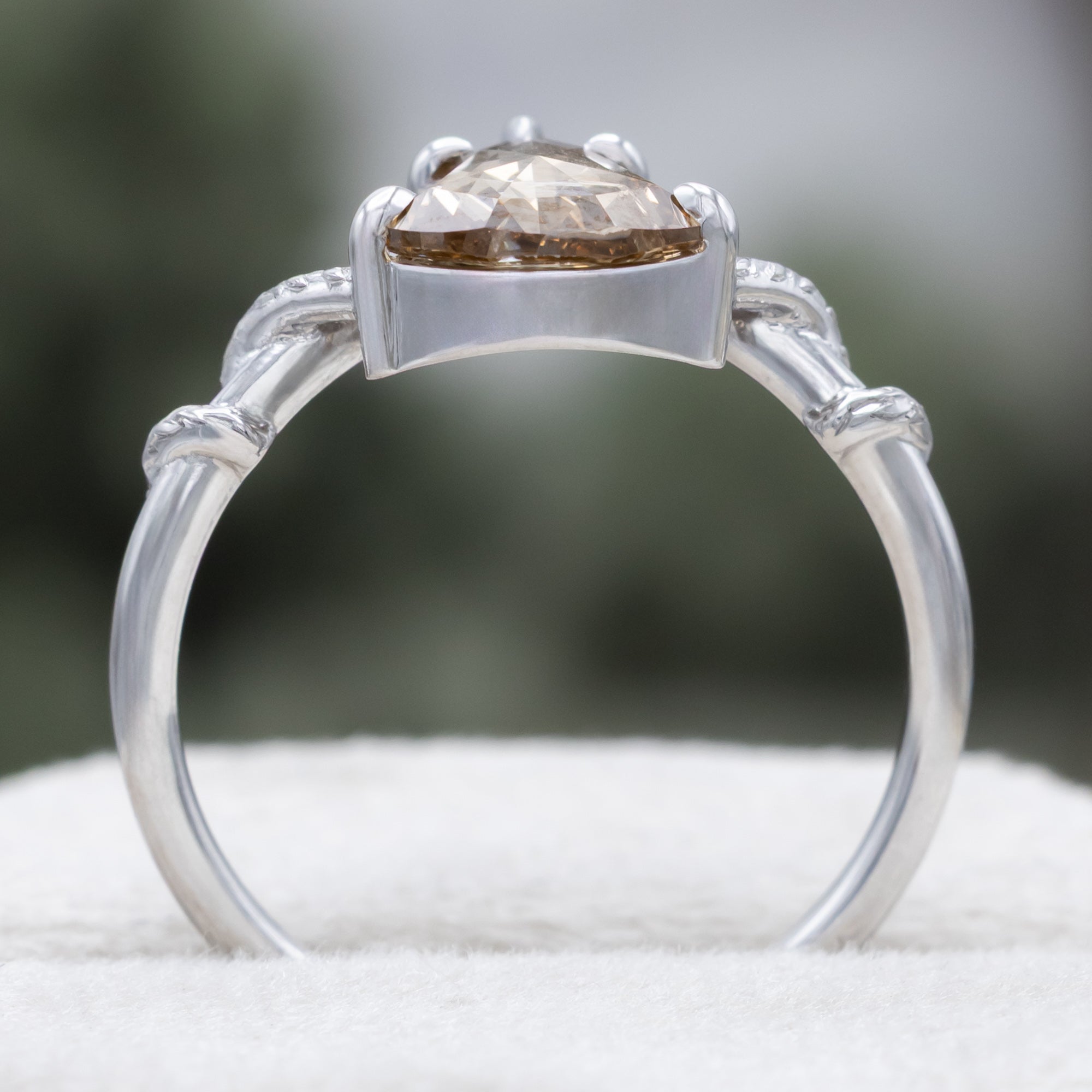 A white gold engagement ring featuring a shield-cut champagne diamond, with vine filigree twisting along the band against a soft blurred green background.