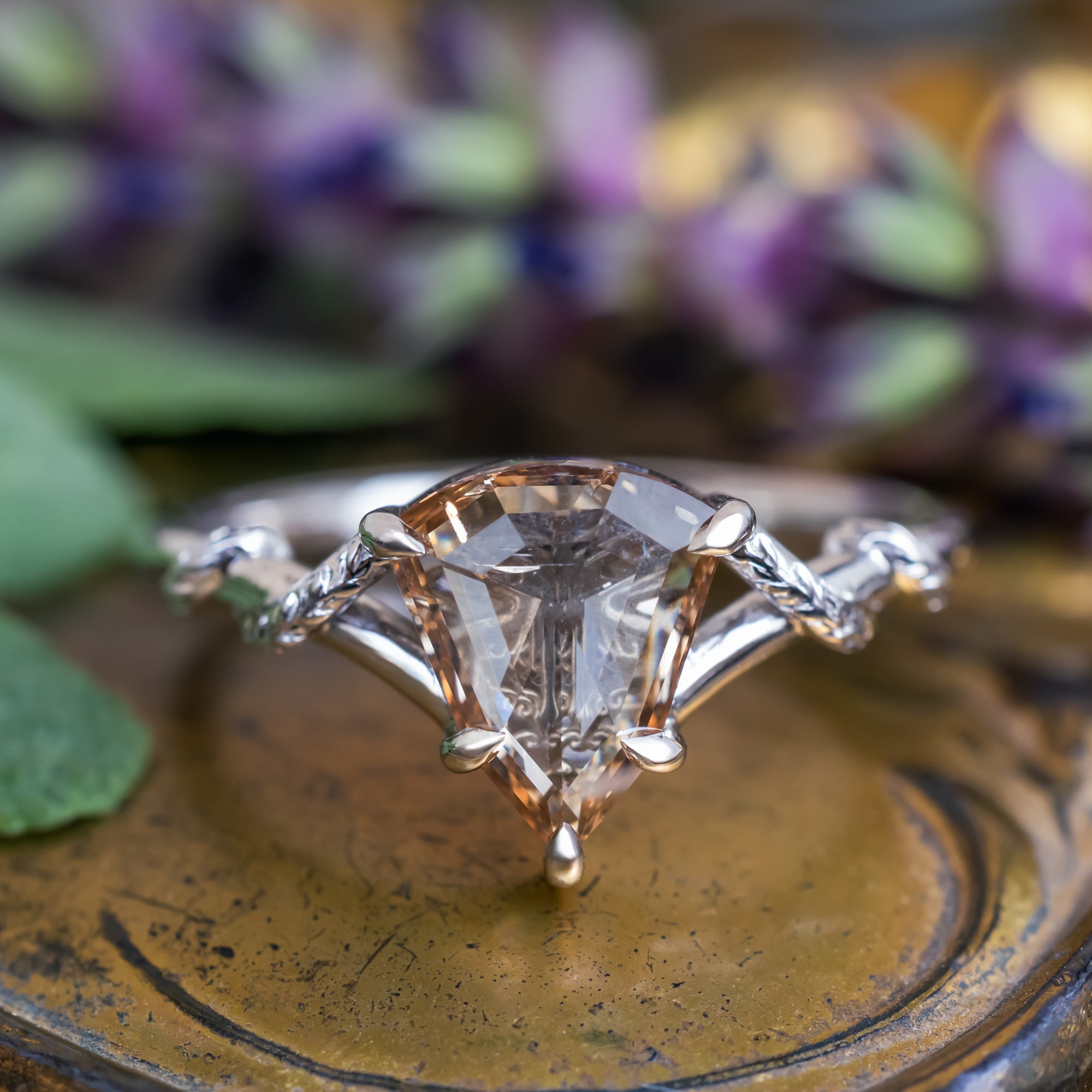A white gold engagement ring featuring a shield-cut champagne diamond, displayed on a textured bronze surface with blurred purple flowers in the background.