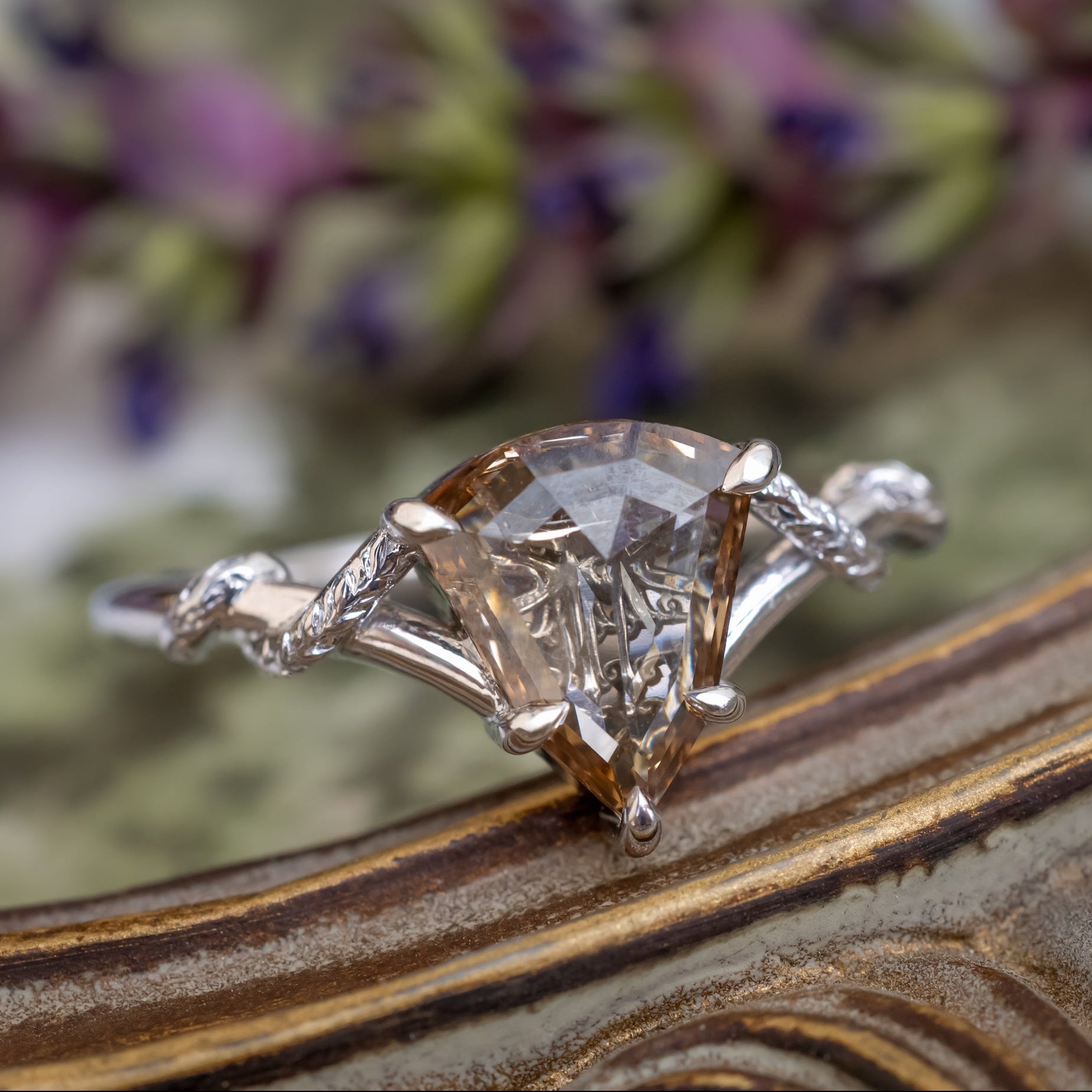 A white gold engagement ring featuring a shield-cut champagne diamond, resting on a textured gold surface with blurred purple flowers in the background.