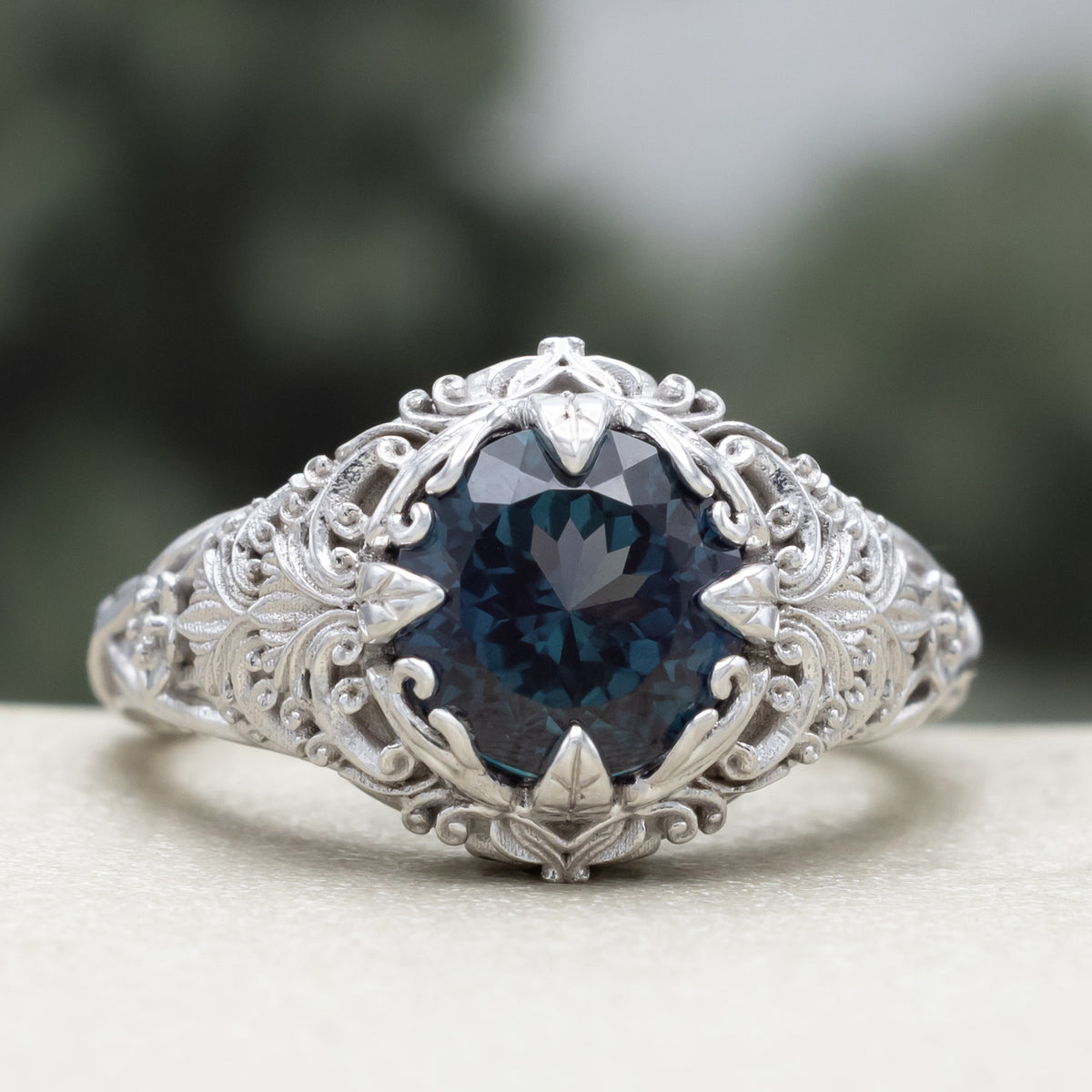 White gold ring with sculptural filigree and round alexandrite center stone against a blurred green background