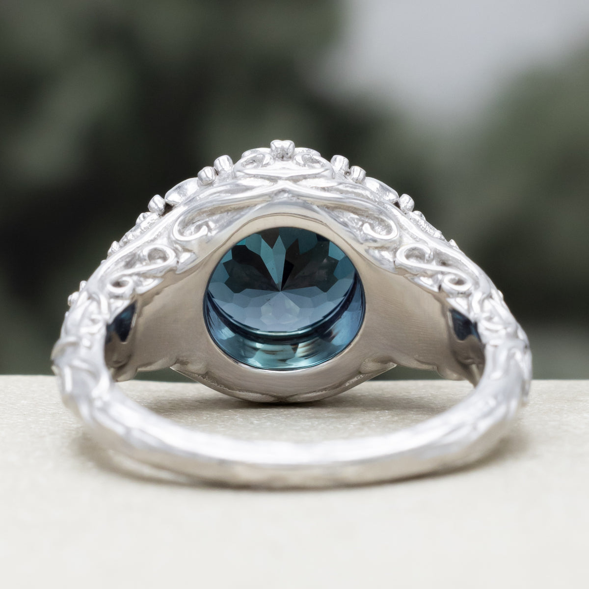 White gold ring with sculptural filigree and round alexandrite center stone against a blurred green background