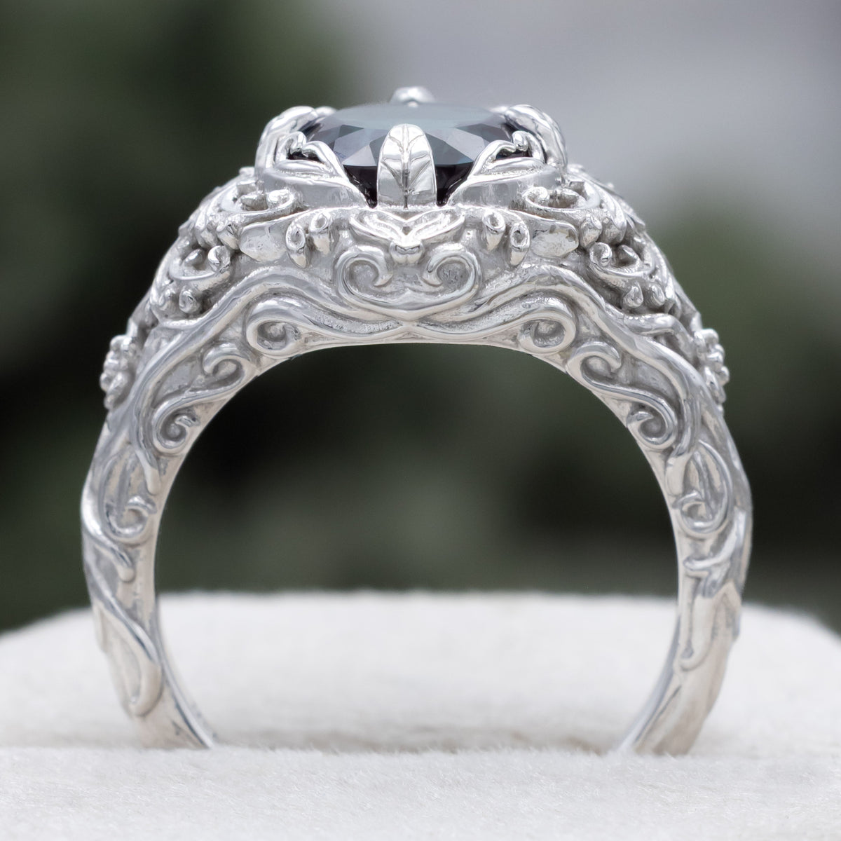White gold ring with sculptural filigree and round alexandrite center stone against a blurred green background